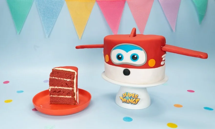 Ein bunter Kinderkuchen in Form eines Super Wings Flugzeugs, mit einer weiteren Kuchenscheibe auf einem kleinen roten Teller daneben, vor einem bunten Partyhintergrund mit Fahnen.