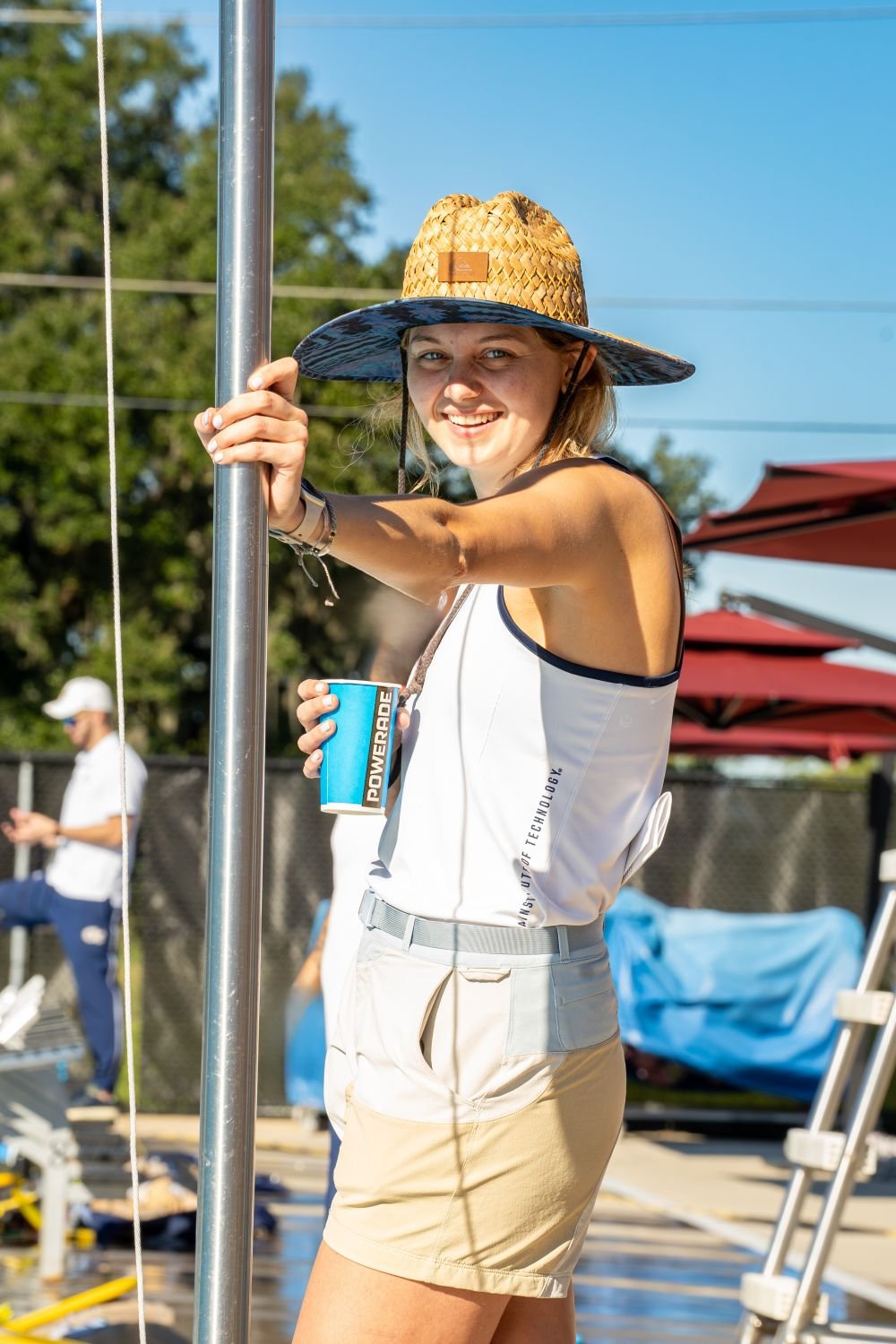 Young woman smiling, wearing a large straw hat and athletic outfit, holding a beverage and leaning on a pole outdoors during sunny weather.