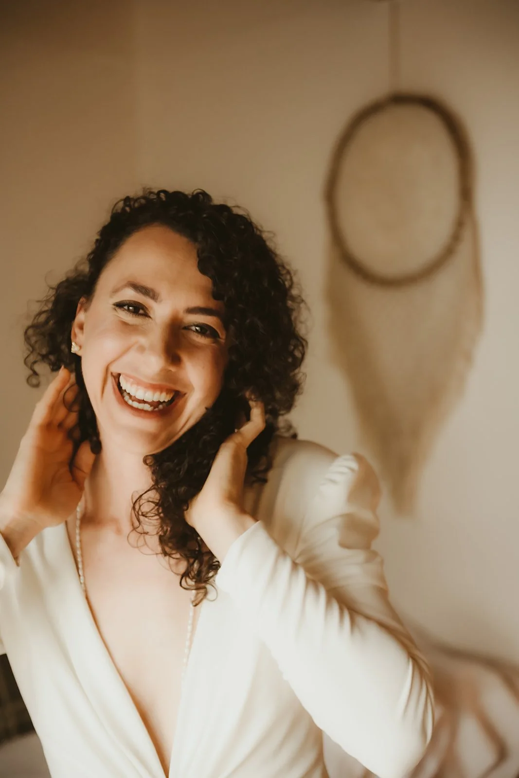 A woman with curly dark hair, wearing a white top and pearl necklace, smiling and holding her neck, standing in front of a wall with a dreamcatcher.