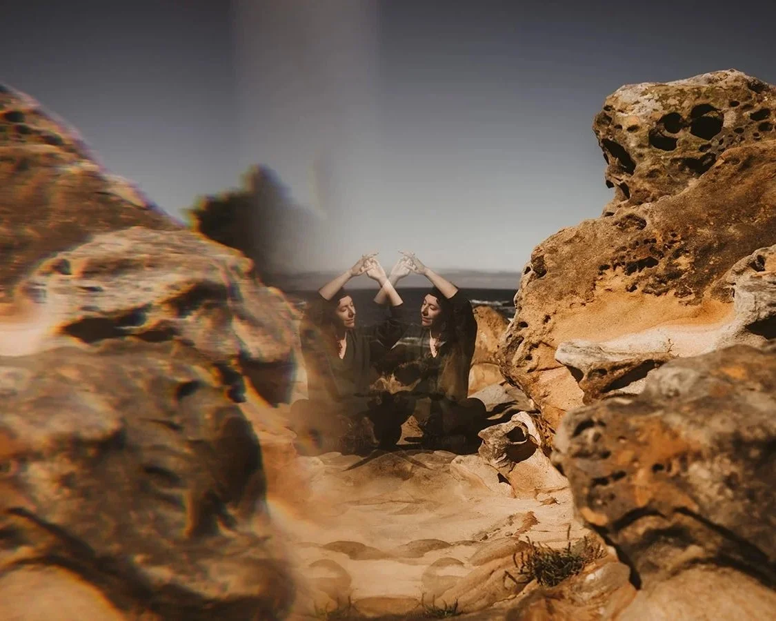 A mirror image of two women sitting and touching hands, with a rocky landscape in the foreground and a desert landscape in the background.