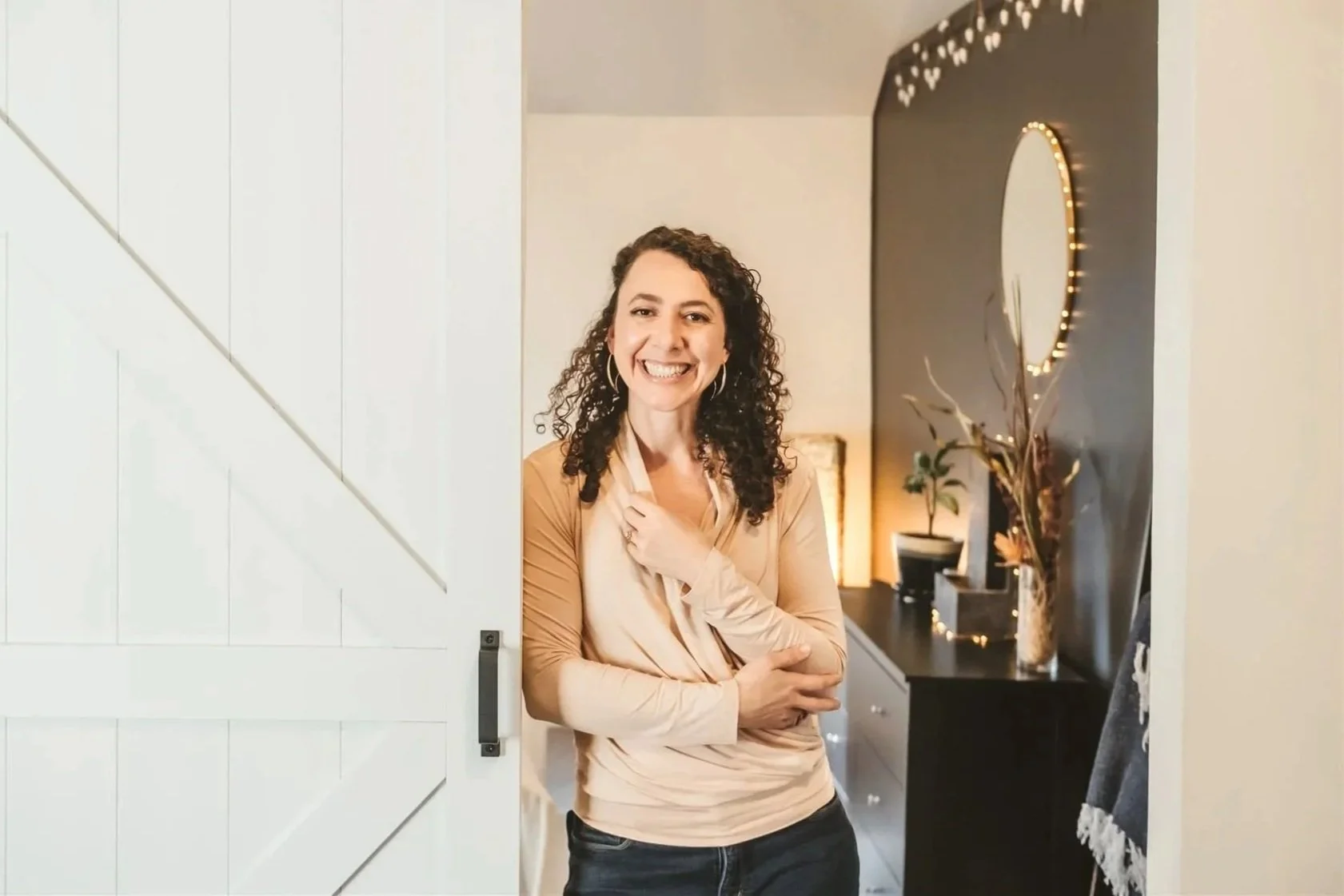 A woman with curly brown hair and hoop earrings smiling in a doorway, with a decorated wall, mirror, and plants in the background.