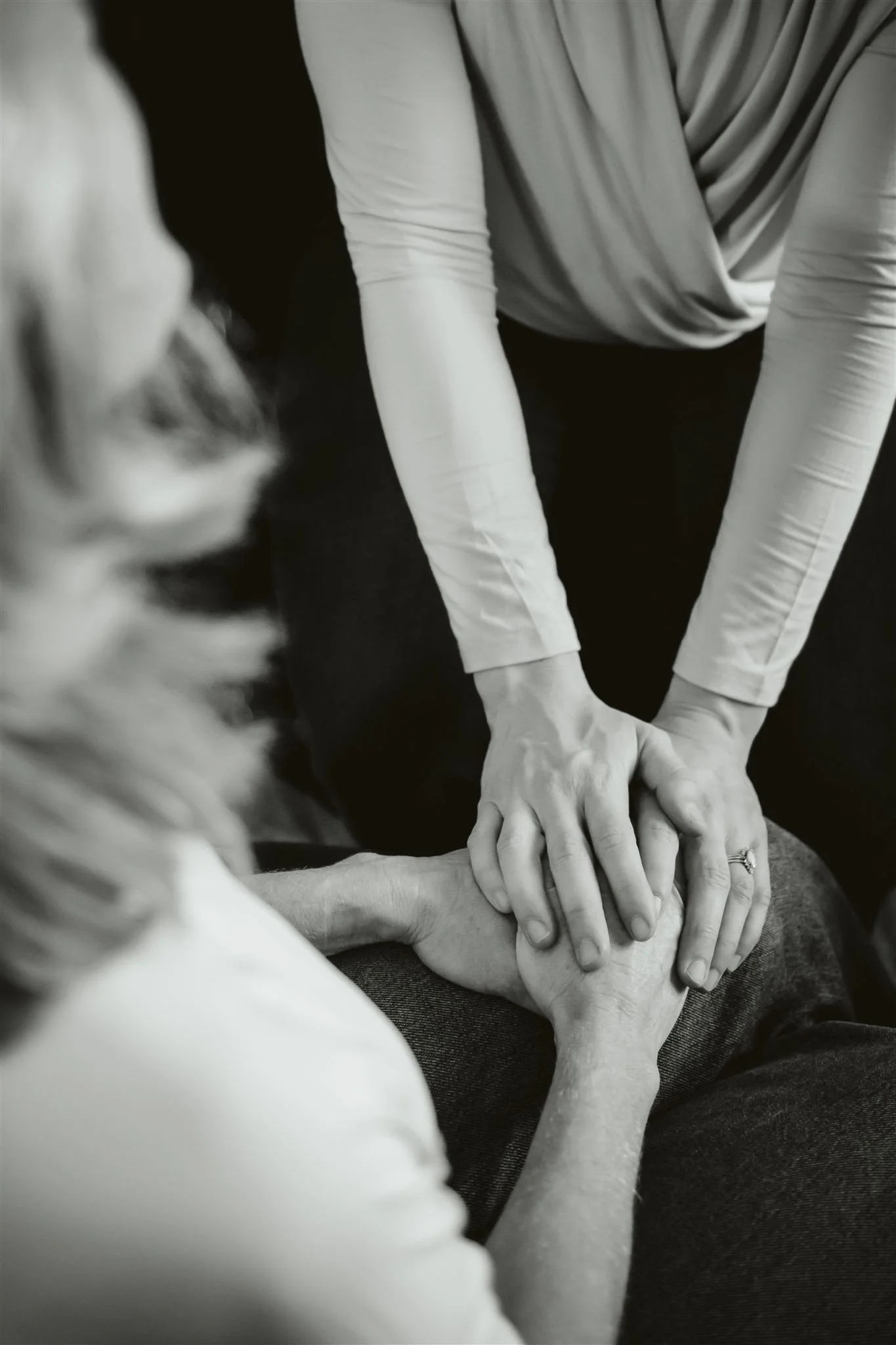 Black and white photo showing a person in a long sleeve shirt with hands resting on another person's knee, while a woman with long hair sits nearby.