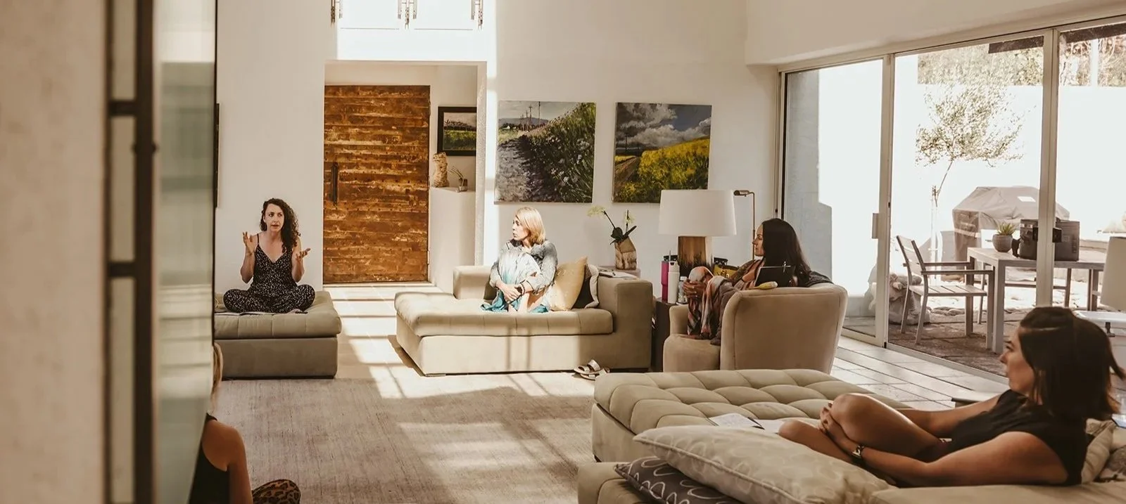 A group of five women gathered in a sunlit living room, sitting on couches and an armchair, engaged in a conversation.