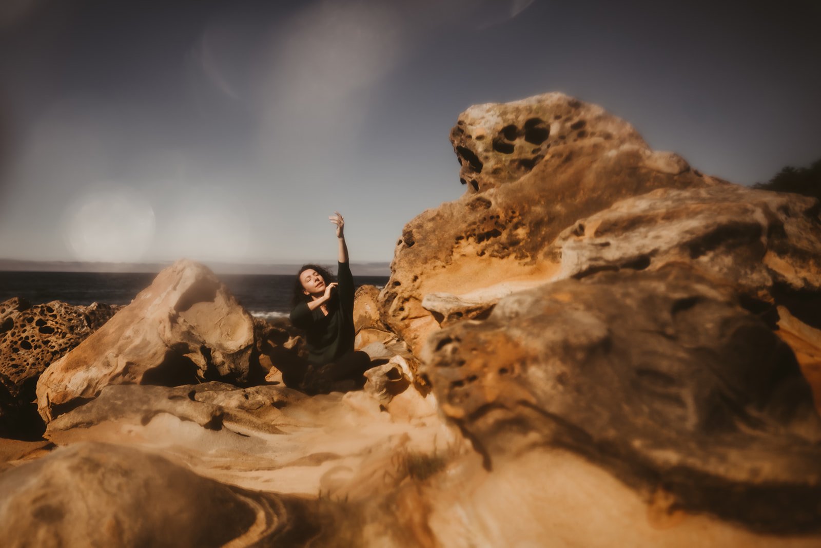 A person sitting among large weathered rocks on a beach, striking a graceful pose with one arm raised, with the ocean and blue sky in the background.
