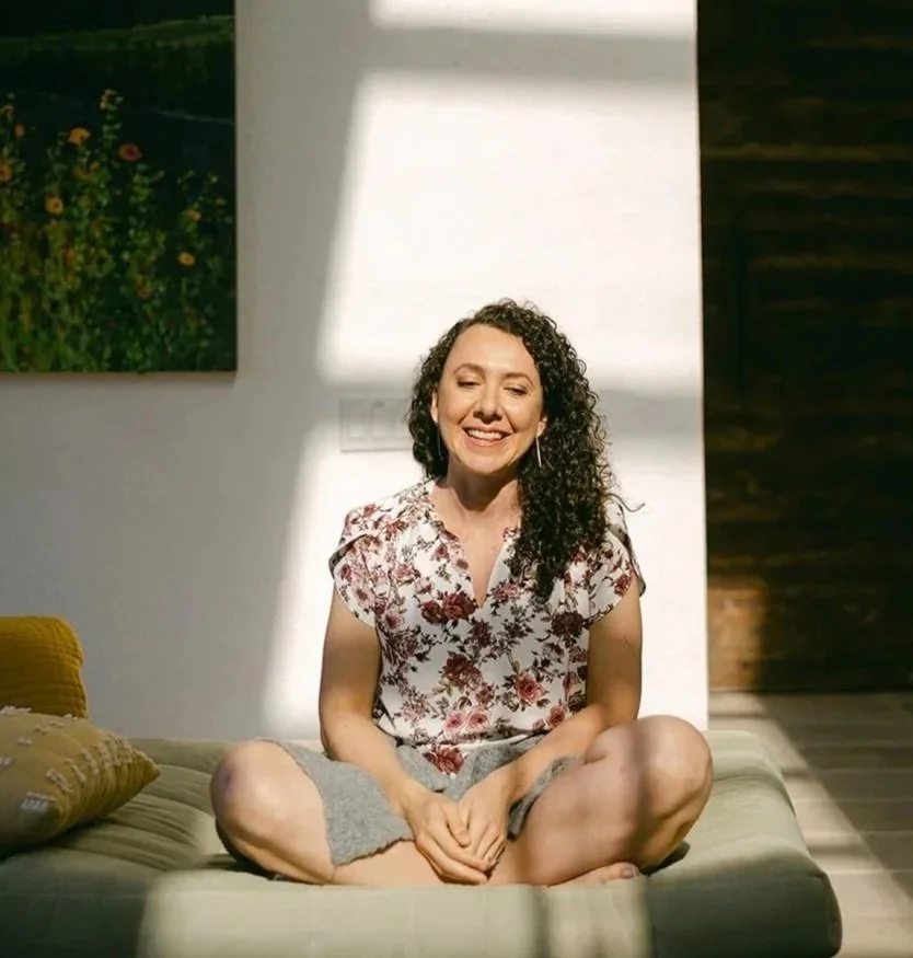 A woman sitting cross-legged on a couch in a sunlit room, smiling, with a floral shirt and gray shorts, with a painting on the wall and a yellow pillow beside her.