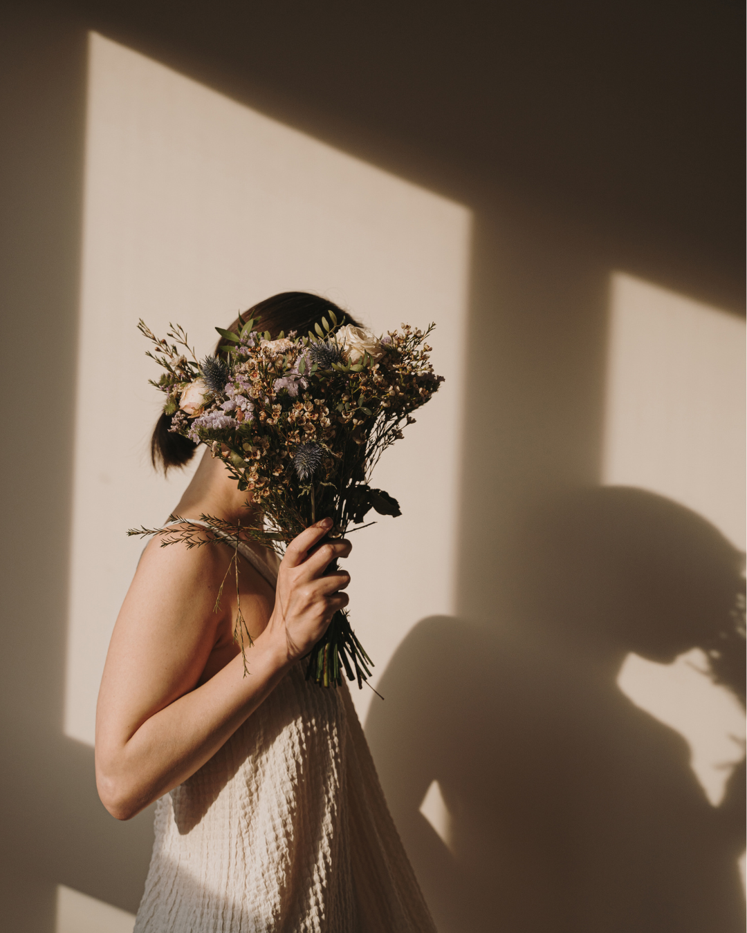 A woman in a beige dress holds a large bouquet of assorted flowers in front of her face, creating a shadow on the wall behind her.