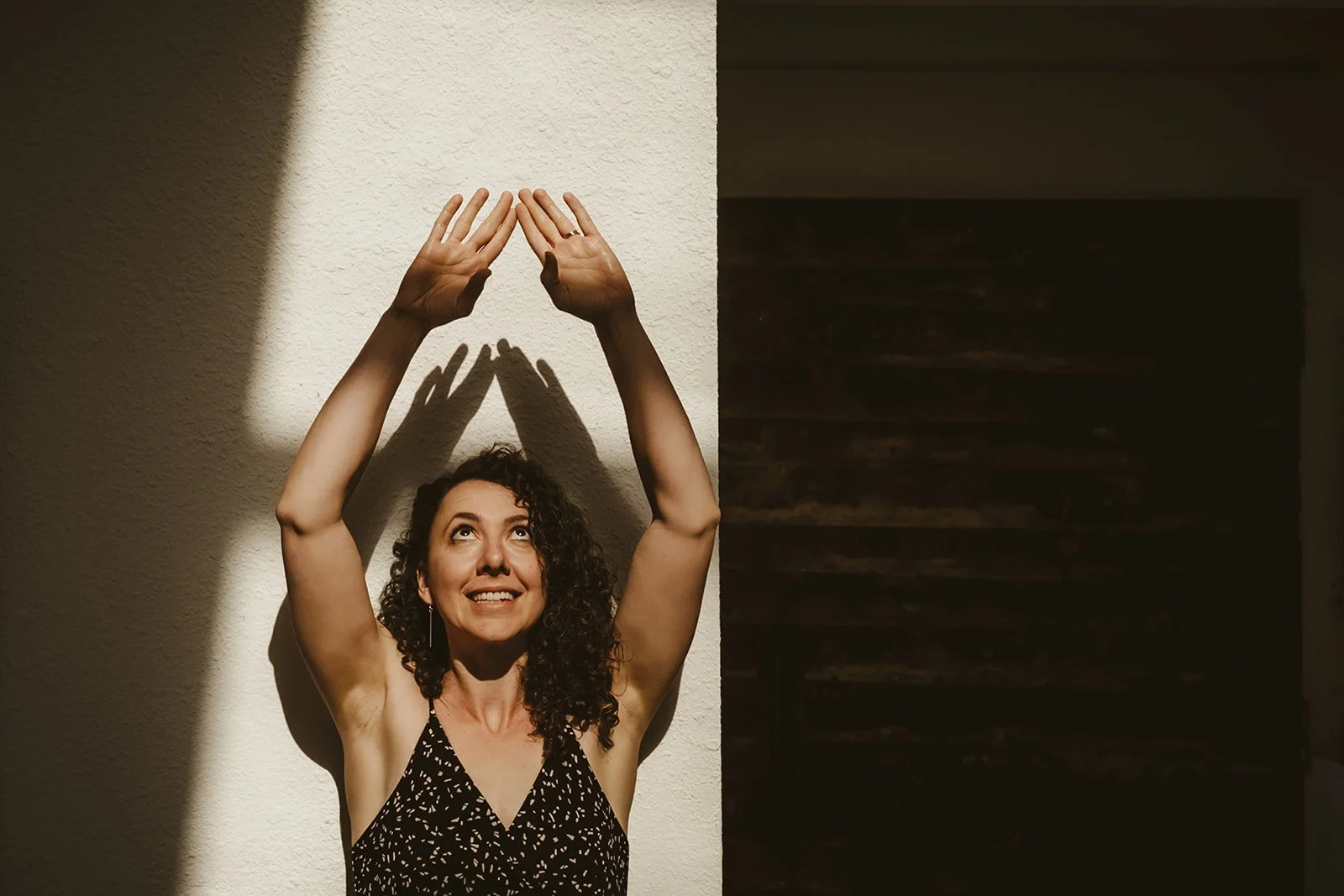 A woman with curly dark hair in a black and white patterned sleeveless top is standing against a wall, raising her arms above her head with her hands touching, creating a shadow of her hands' reflection on the wall.