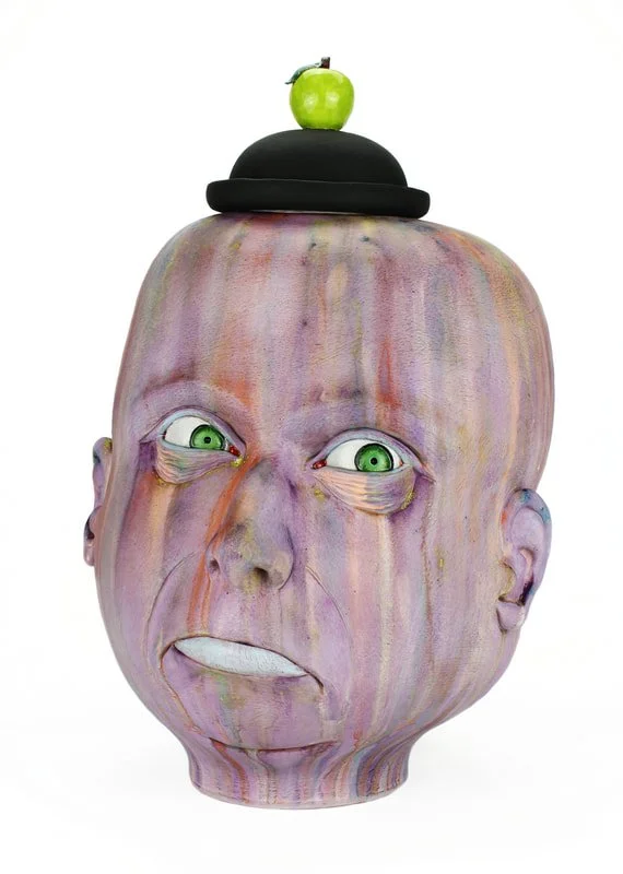 A sculpted ceramic vessel resembling a human head with exaggerated facial features, painted in pastel tones. It has green eyes, a protruding tongue, and wears a black hat with a green apple on top.