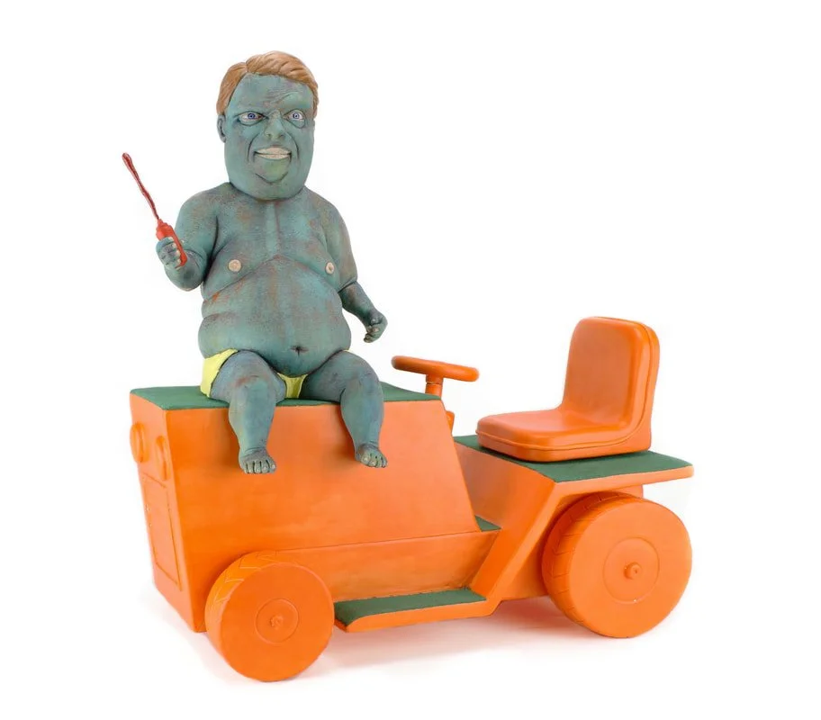 A humorous sculpture of a baby with a human face, green body, and yellow diaper, sitting on an orange toy vehicle with an orange seat and large orange wheels, holding a small red stick.