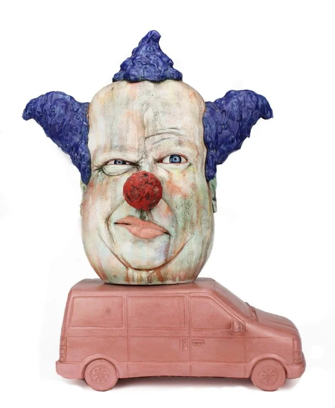 A sculpture of a clown's face with blue hair, a red nose, and a facial expression of confusion or scorn, mounted on a pink toy car base.