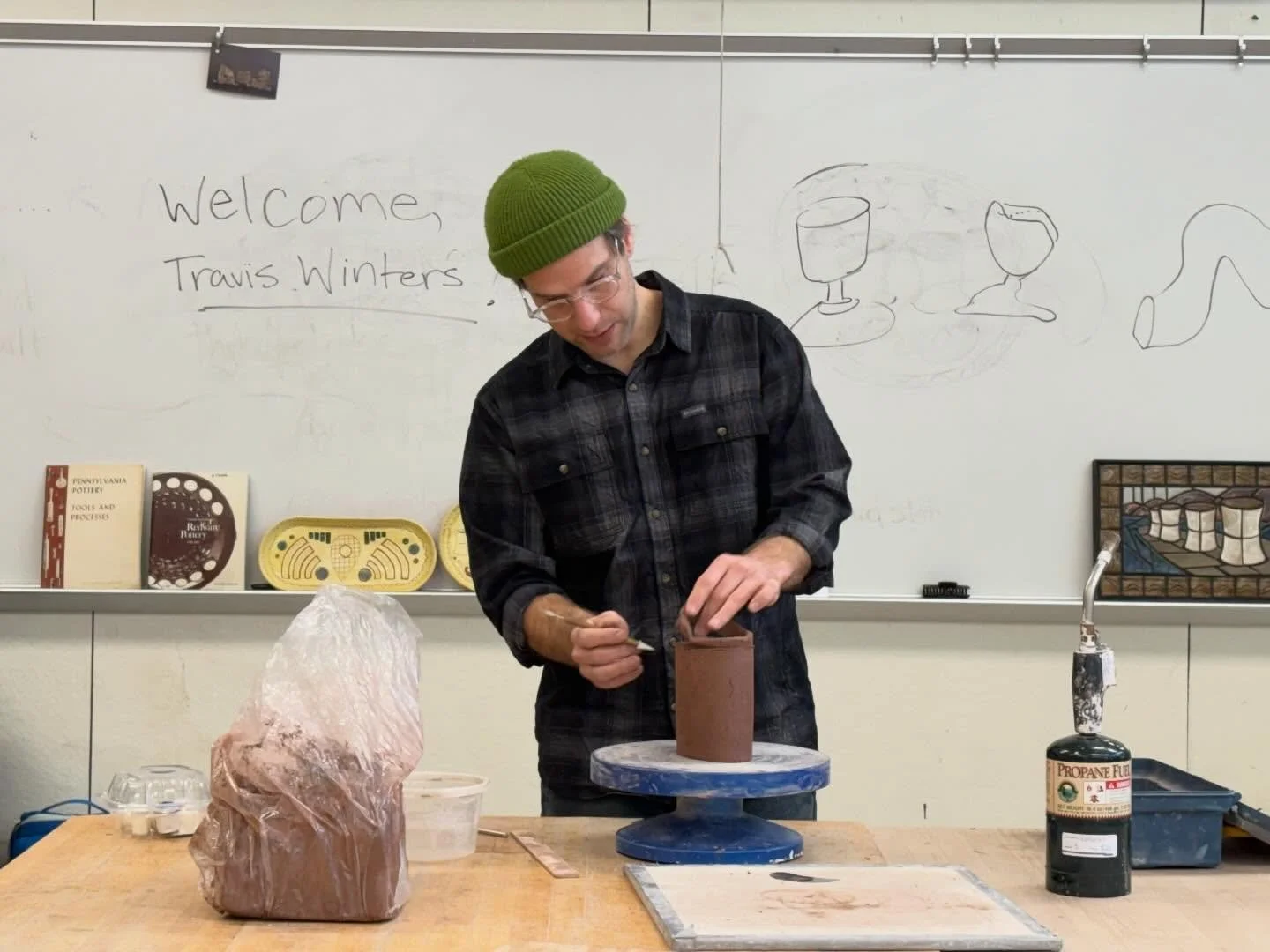 A man wearing glasses, a green beanie, and a black plaid shirt is working on a pottery piece on a potter's wheel in a classroom. Behind him, a whiteboard has the message 'Welcome, Travis Winters' and some sketches of glasses and other objects. There are books, a large rock of clay, and a propane torch on the table.