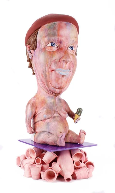 A surreal sculpture of a baby with an oversized head featuring a human face, sitting on a platform made of pink clay pipes, holding a small object in one hand.