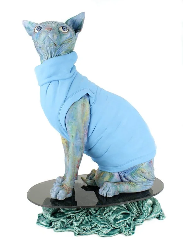 A sculpture of a colorful hairless cat with large eyes, wearing a light blue shirt, sitting on a black oval platform with crumpled green fabric underneath.