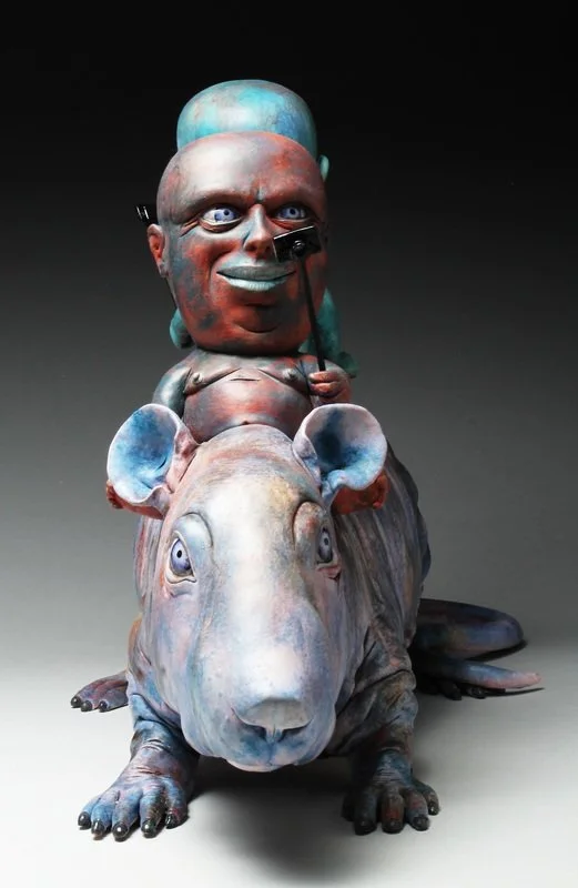 A colorful sculpture of a baby riding a large rat, with the baby holding a stick and wearing a hat, against a dark background.