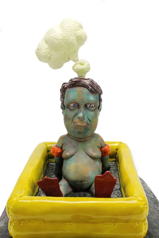 A colorful sculpture of a person with a greenish skin tone sitting in a yellow bathtub. A piece of cauliflower hovers above their head, emitting greenish fumes.