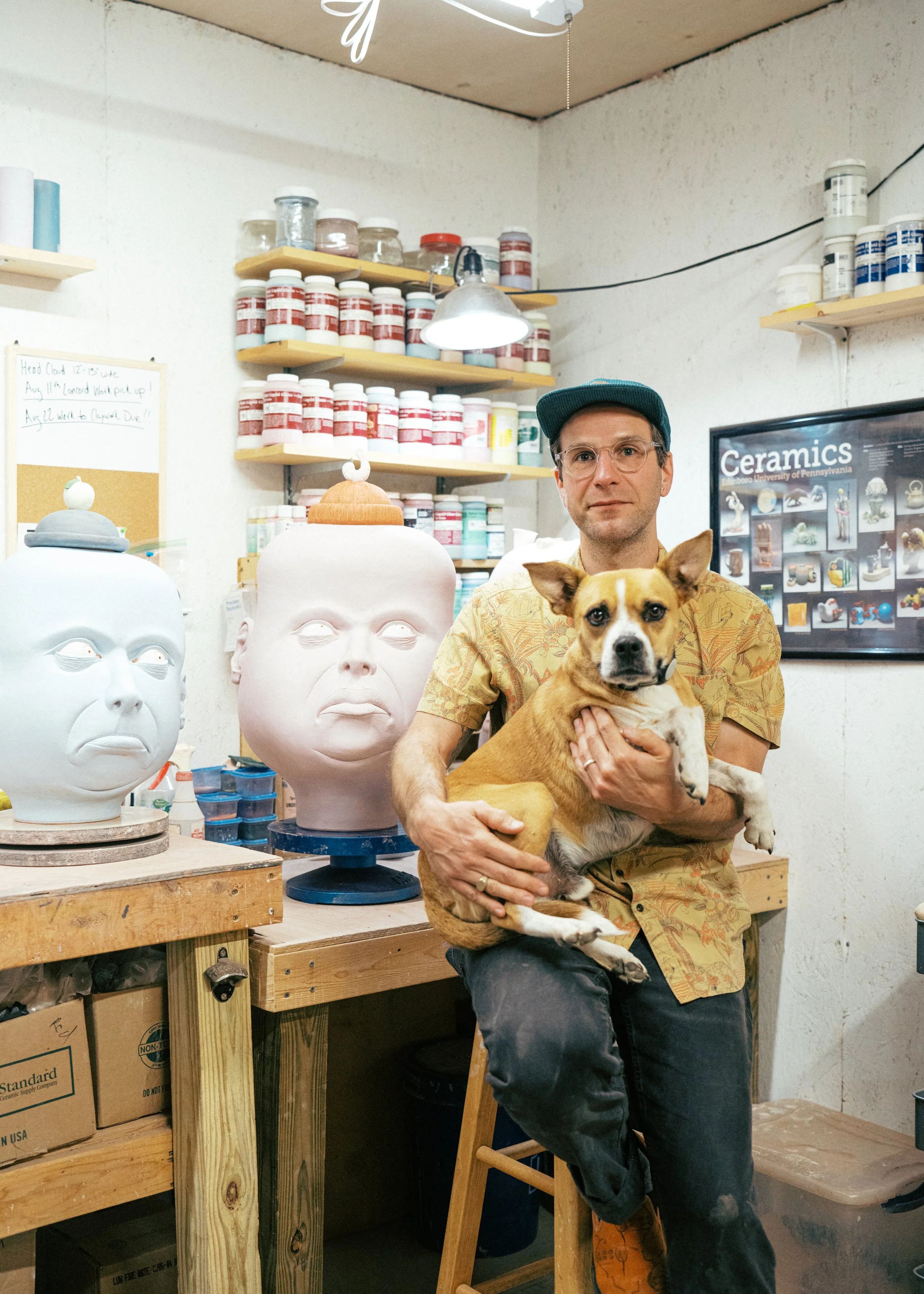 A man holding a tan and white dog stands in a ceramics studio, with sculpted head busts and shelves of paint jars around him. The studio has a wooden worktable and a framed ceramics poster on the wall.
