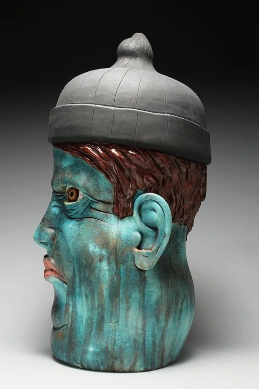 A sculpture of a human head with a gray beanie hat, featuring a detailed face with blue-green skin, brown hair, and an exaggerated facial expression.