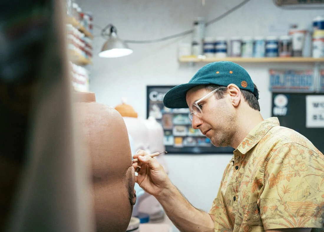 A man wearing glasses, a blue cap, and a yellow Hawaiian shirt is painting the nipple of a large, pink sculpture or model in an art studio or workshop.