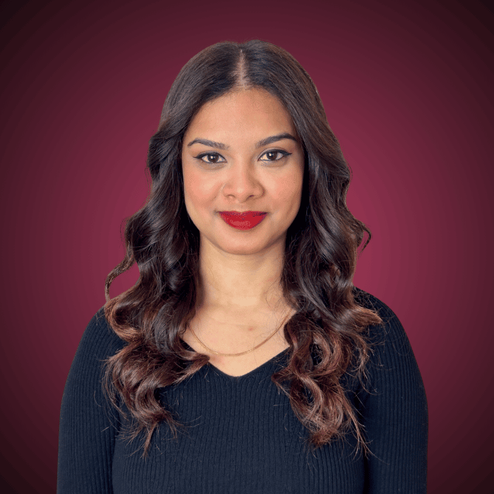 A woman with long, wavy brown hair, wearing a black top, posing against a maroon background.