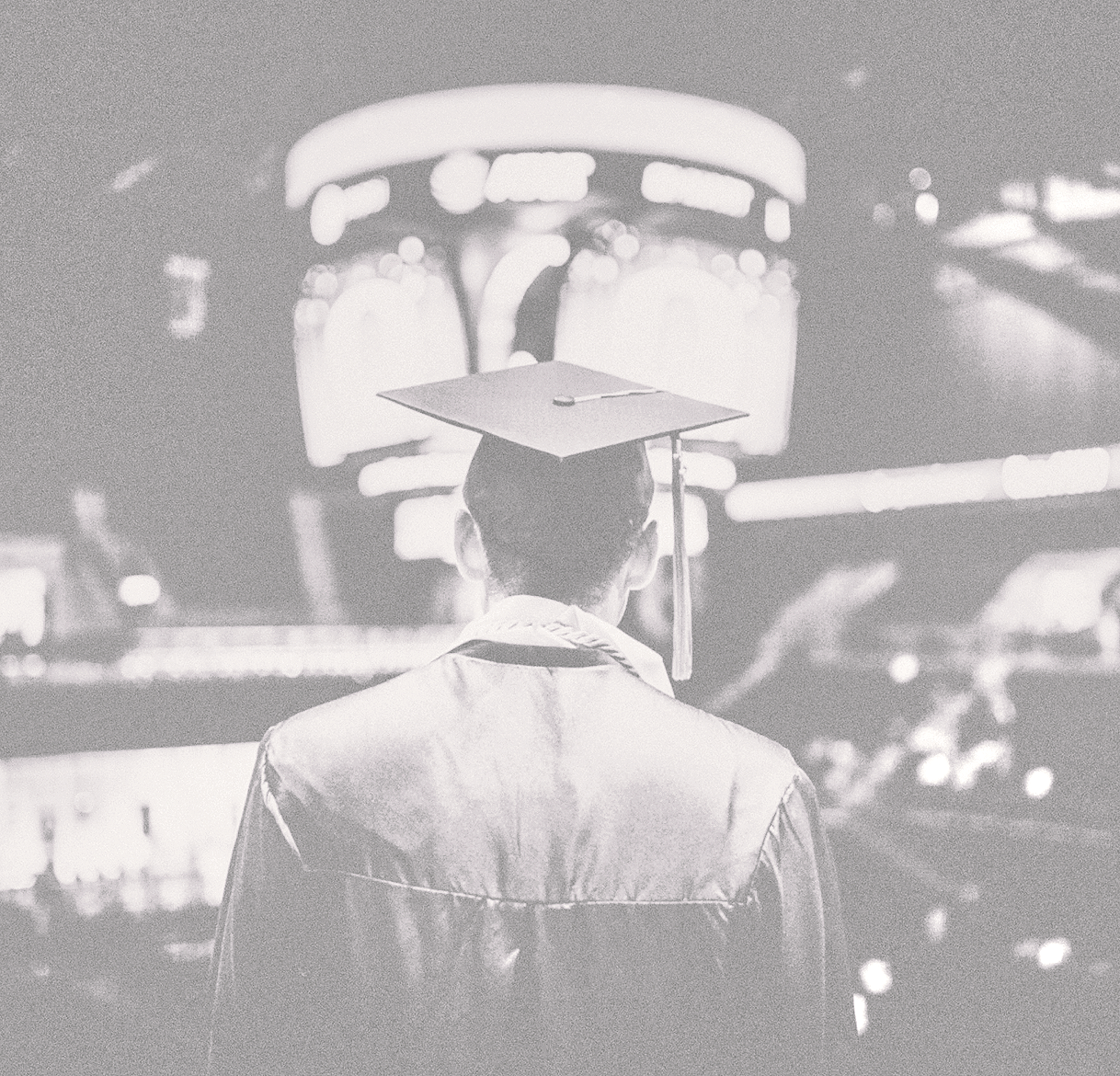 A person wearing a graduation cap and gown facing away in an outdoor night scene with illuminated signs and blurred lights in the background.