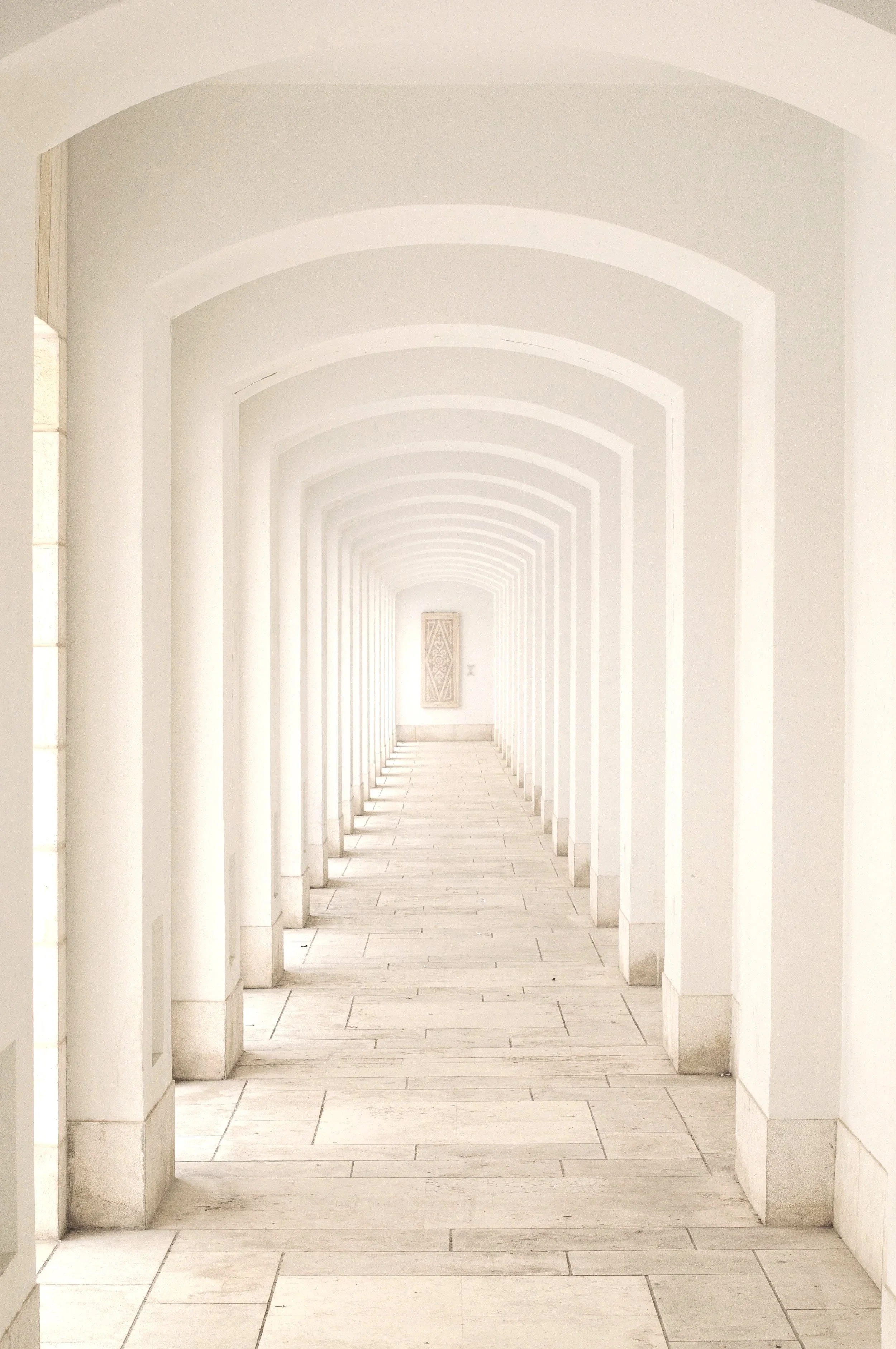 A white corridor with arched doorways and a decorative panel at the end, lined with tiled flooring.