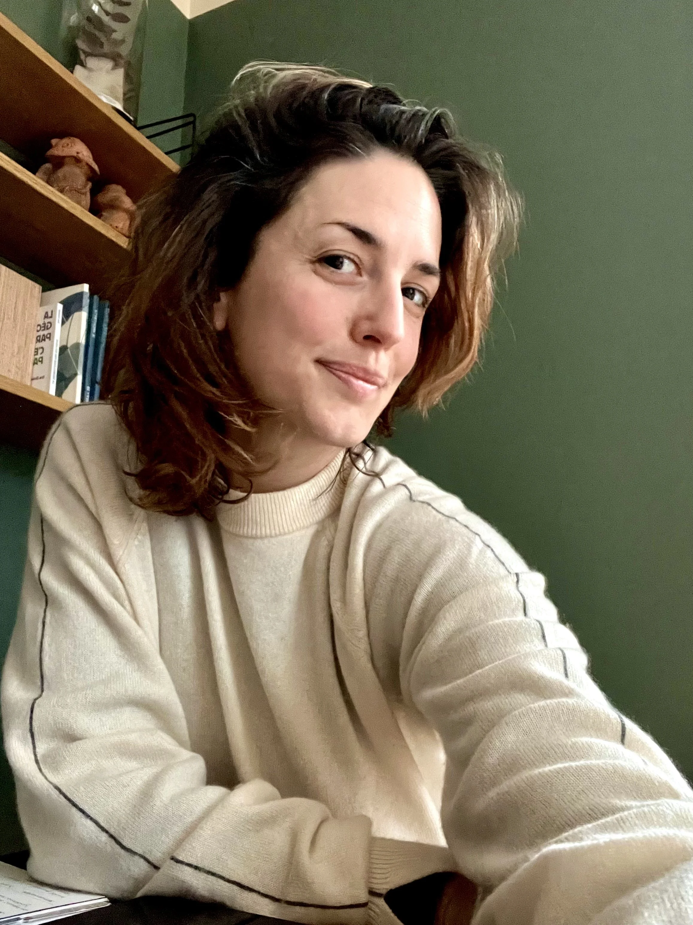Une femme aux cheveux bruns bouclés, souriante, portant un sweatshirt beige, assise devant un mur vert avec une étagère en bois contenant des livres et des objets décoratifs.
