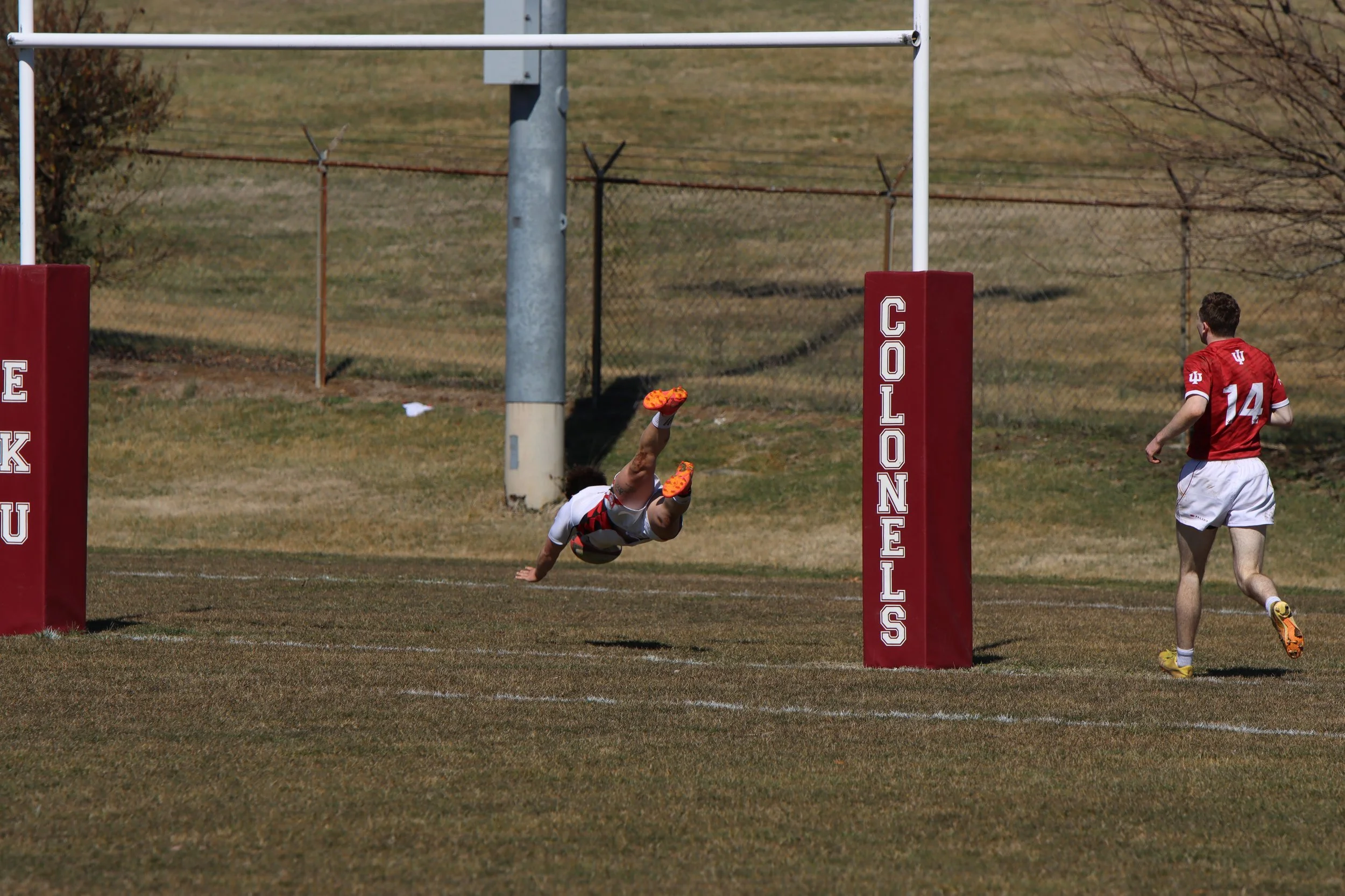 A rugby player in red and white uniform is diving towards the goal post, with another player nearby on the field during daytime.