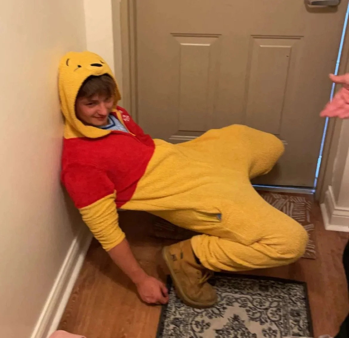 A person dressed in a Winnie the Pooh costume, sitting on the floor and leaning against the wall with a smile.