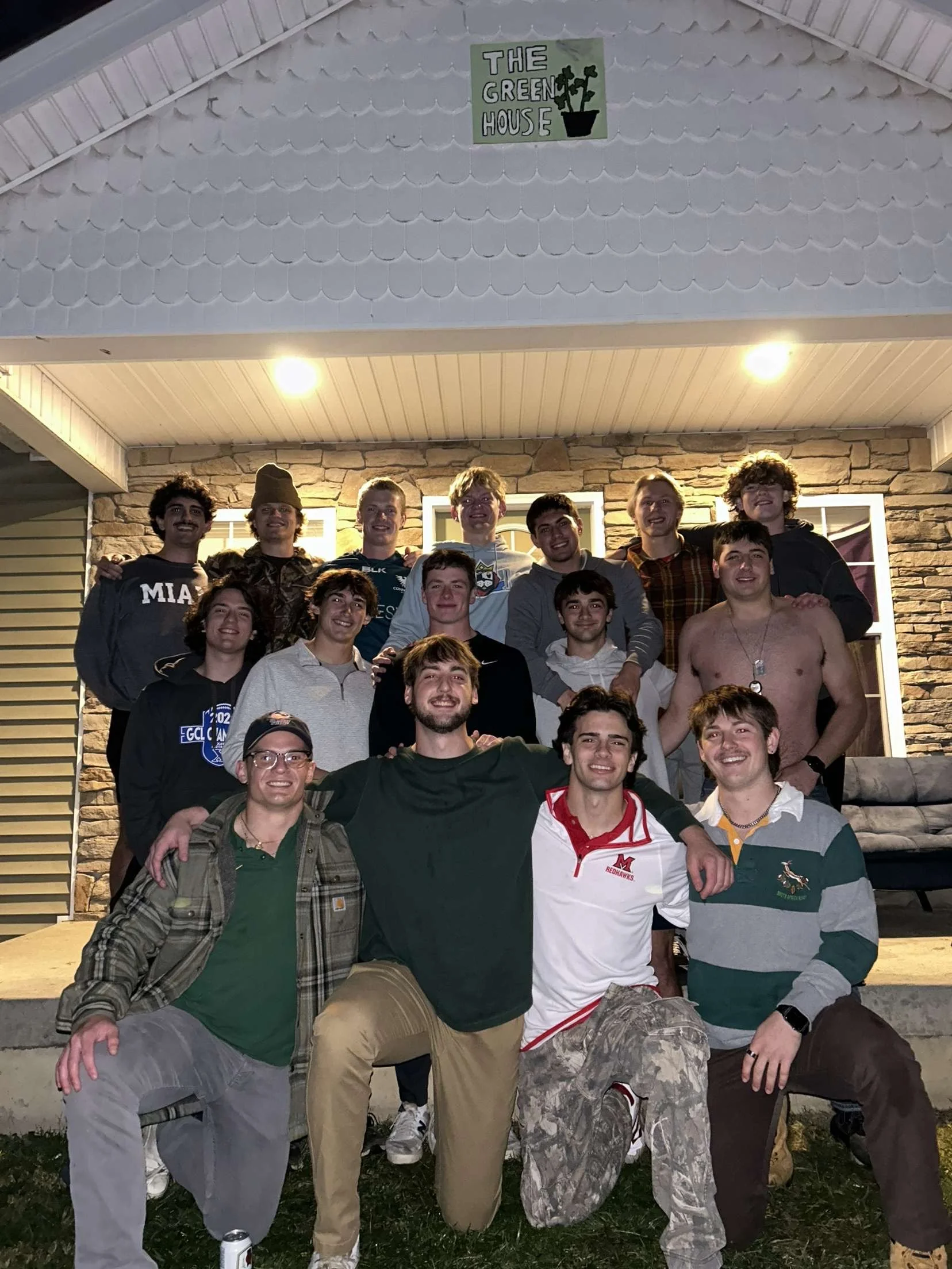 A group of young men posing outside a house at night, smiling and enjoying each other's company. They are standing on a grassy area in front of a stone house with large windows, some wearing casual clothes, and a few shirtless, with a night sky overhead.