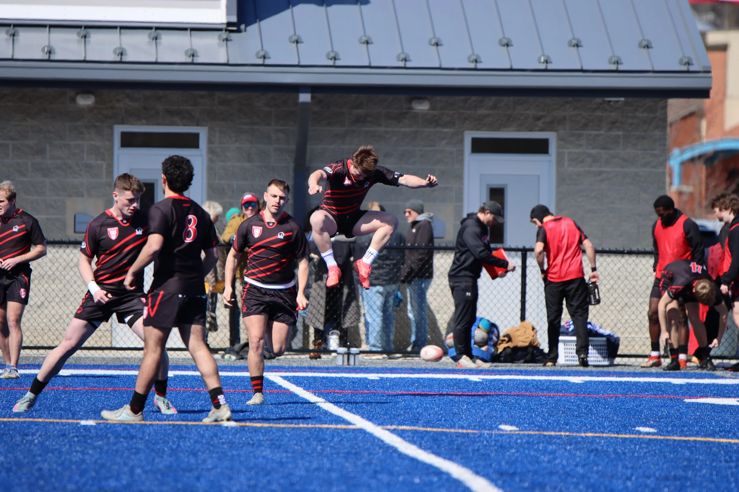 A rugby player jumps to catch or block the ball in a match with teammates and opponents on a blue turf field, with a building and spectators in the background.