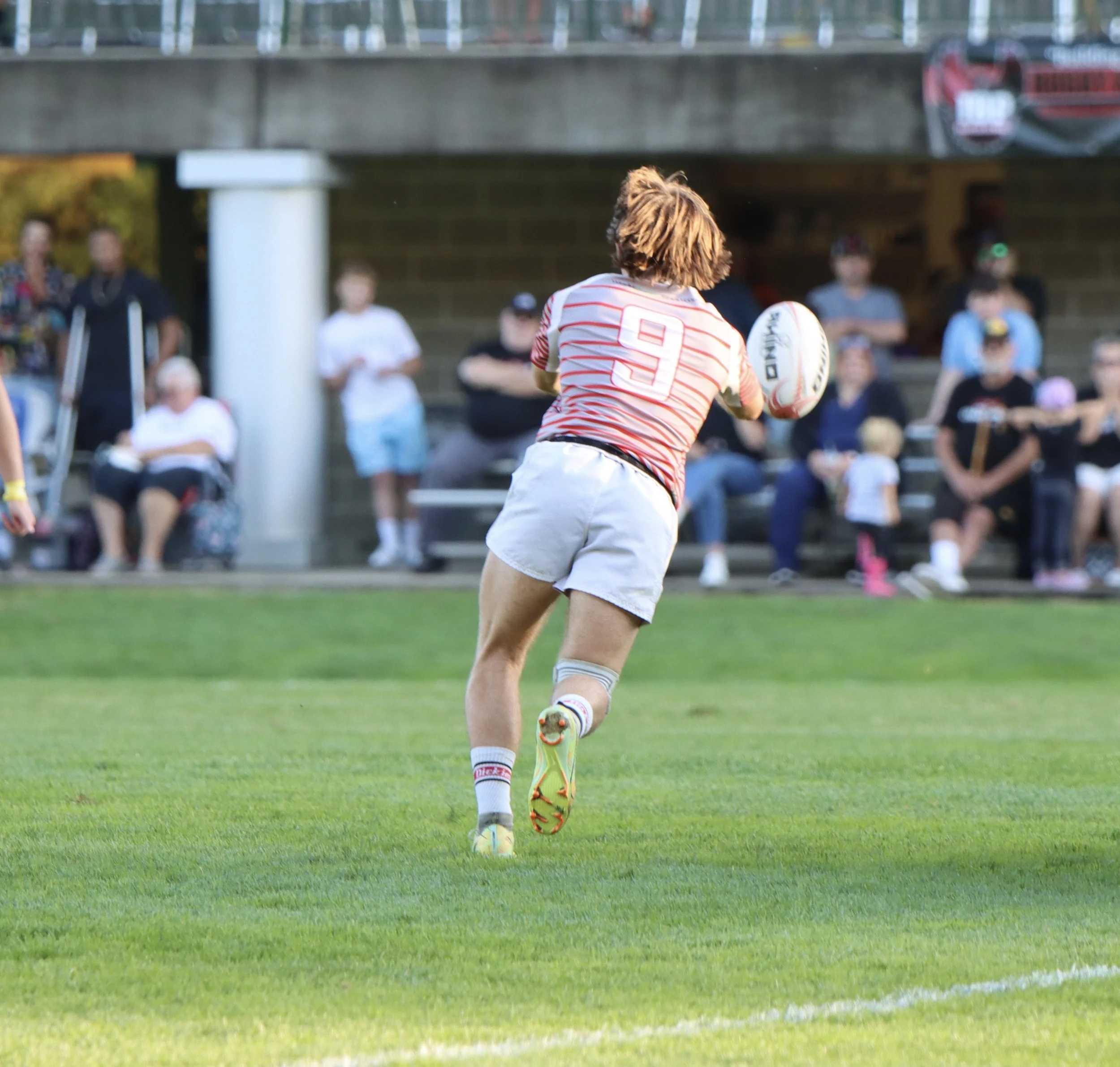 A female rugby player with short curly hair, wearing a red and white striped jersey with the number 9, white shorts, and colorful cleats, is running on a green field while holding a rugby ball. Spectators are sitting and standing on benches in the background under a bridge.