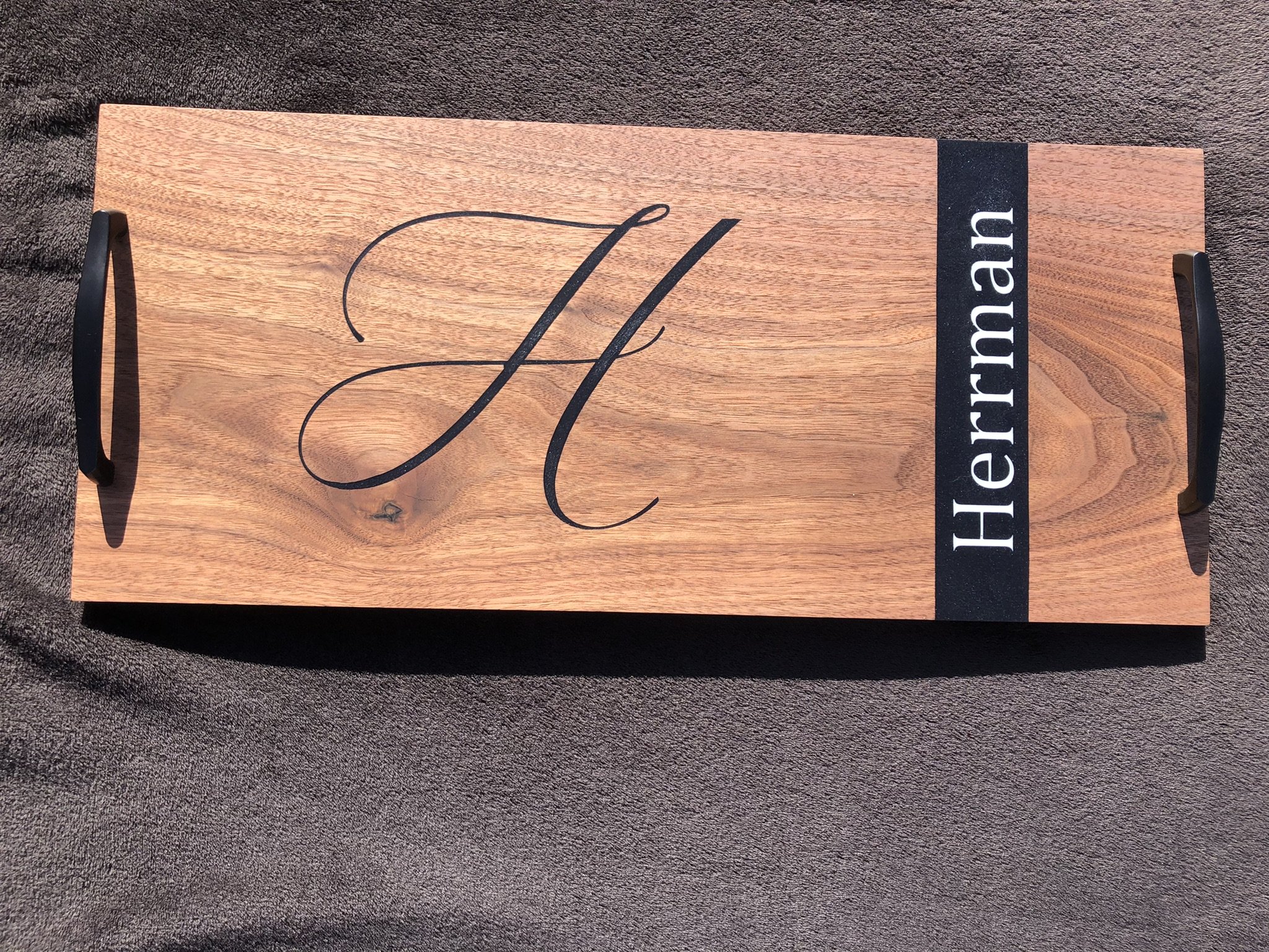 A wooden serving tray with black handles, featuring a large calligraphic letter 'H' and a black band with the word 'Herrmann' written on it.