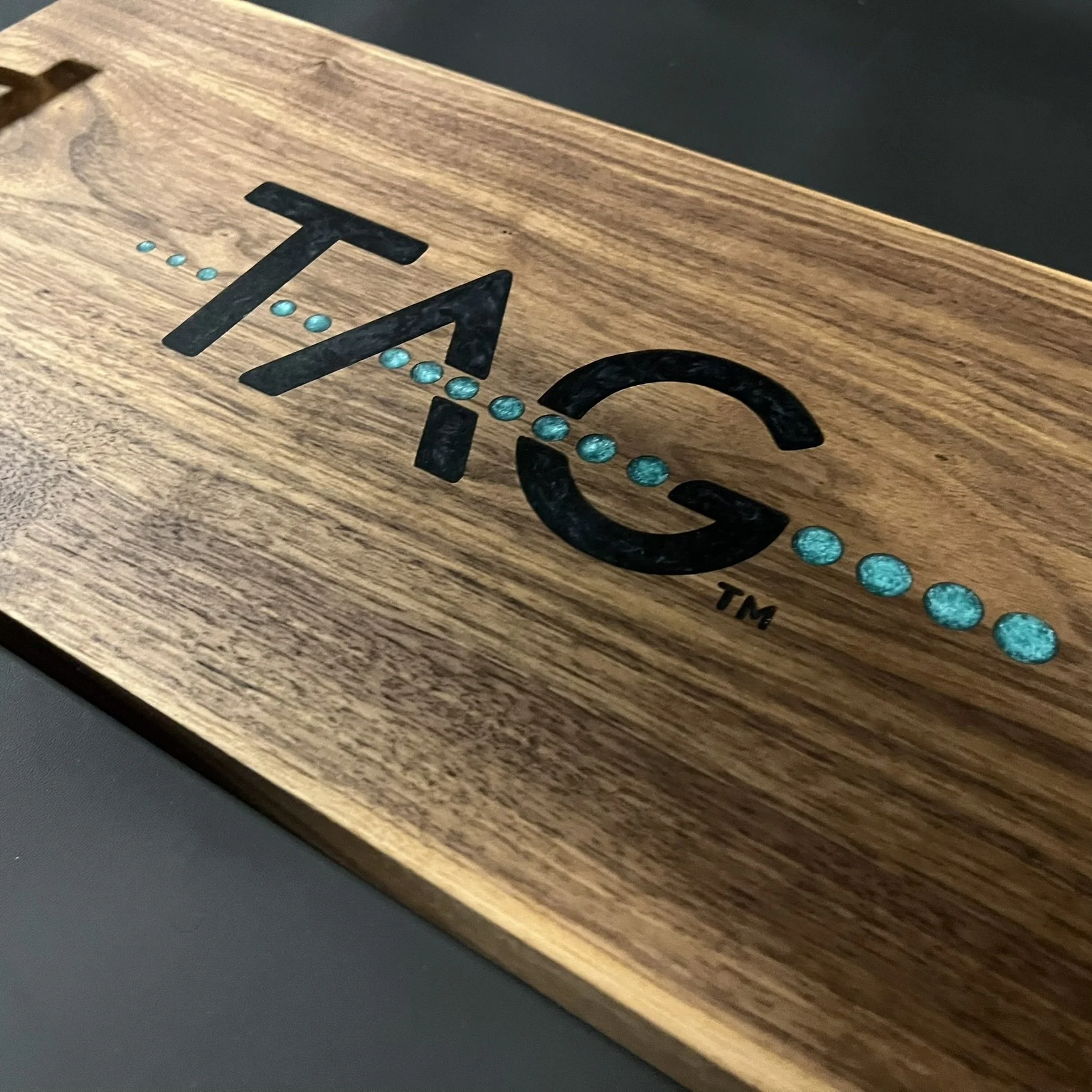 Close-up view of a wooden surface with the TMSC logo engraved, featuring black letters and blue dot accents.
