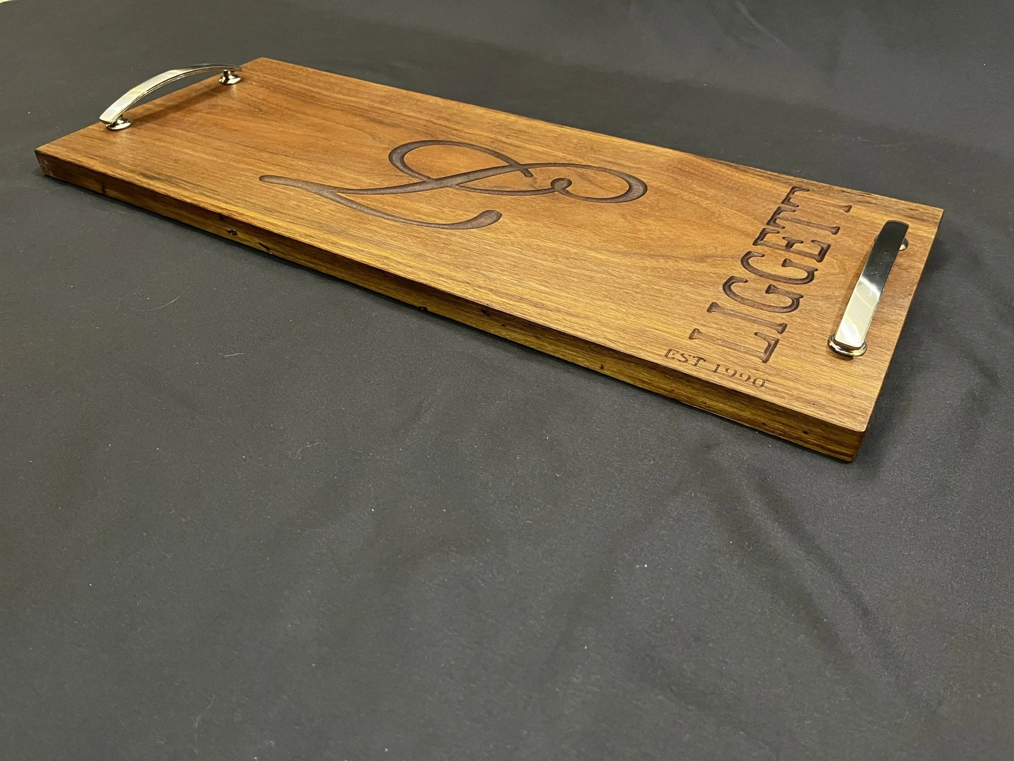 A rectangular wooden board with the word "Legget" and the letter "L" engraved on it, metal handles on each end, and the words "EST 1940" engraved at the bottom right corner.