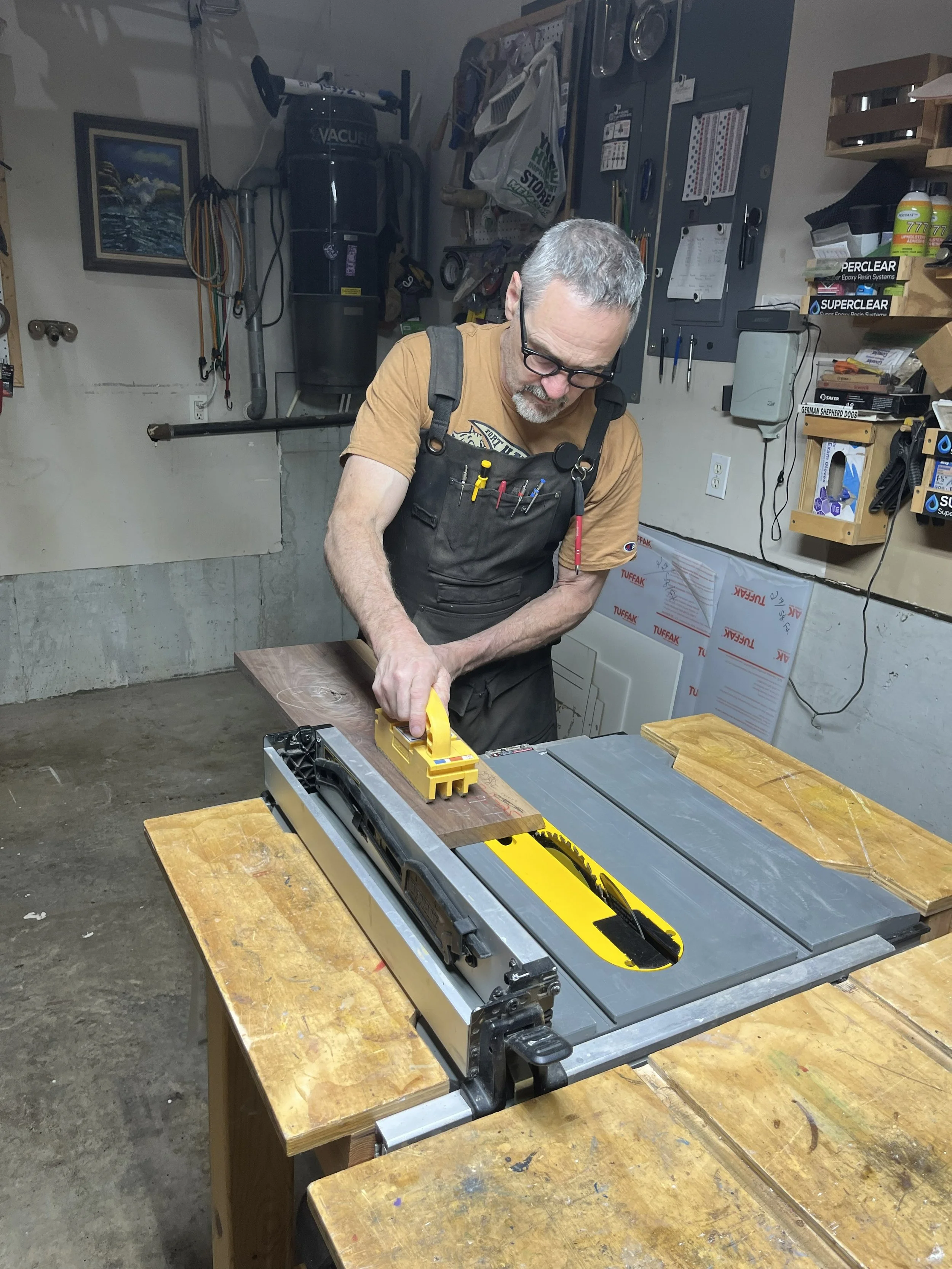 A man with gray hair, glasses, and a beard wearing a brown T-shirt and black work apron is using a yellow hand planer on a piece of wood at a woodworking workbench in a garage workshop.