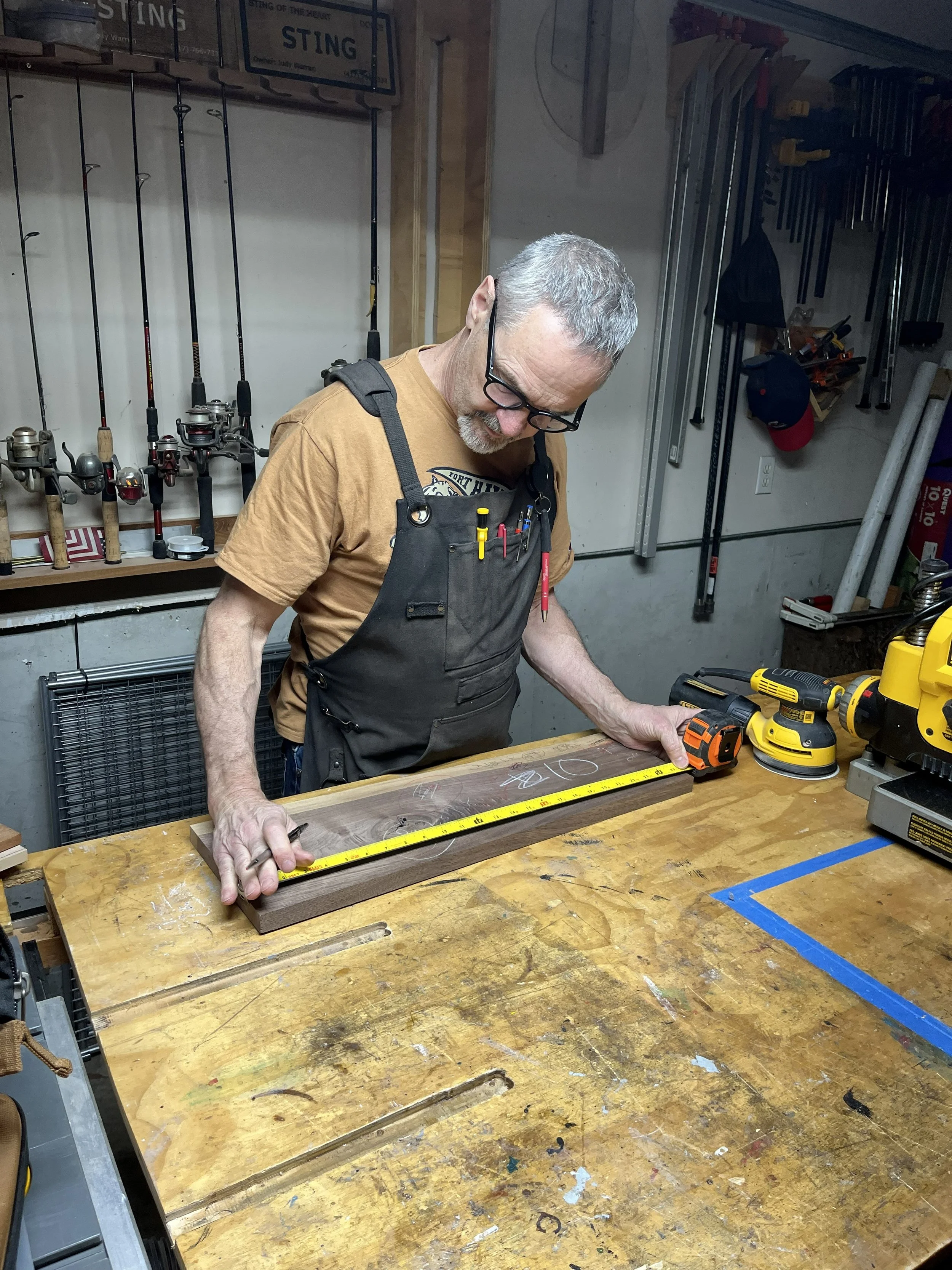 An older man with gray hair, glasses, and a beard is measuring a piece of wood on a workbench in a woodworking shop. The man is wearing a brown T-shirt and a black apron with tools in the pocket. The shop has various tools, including power sanders and clamps, and fishing rods hanging on the wall in the background.