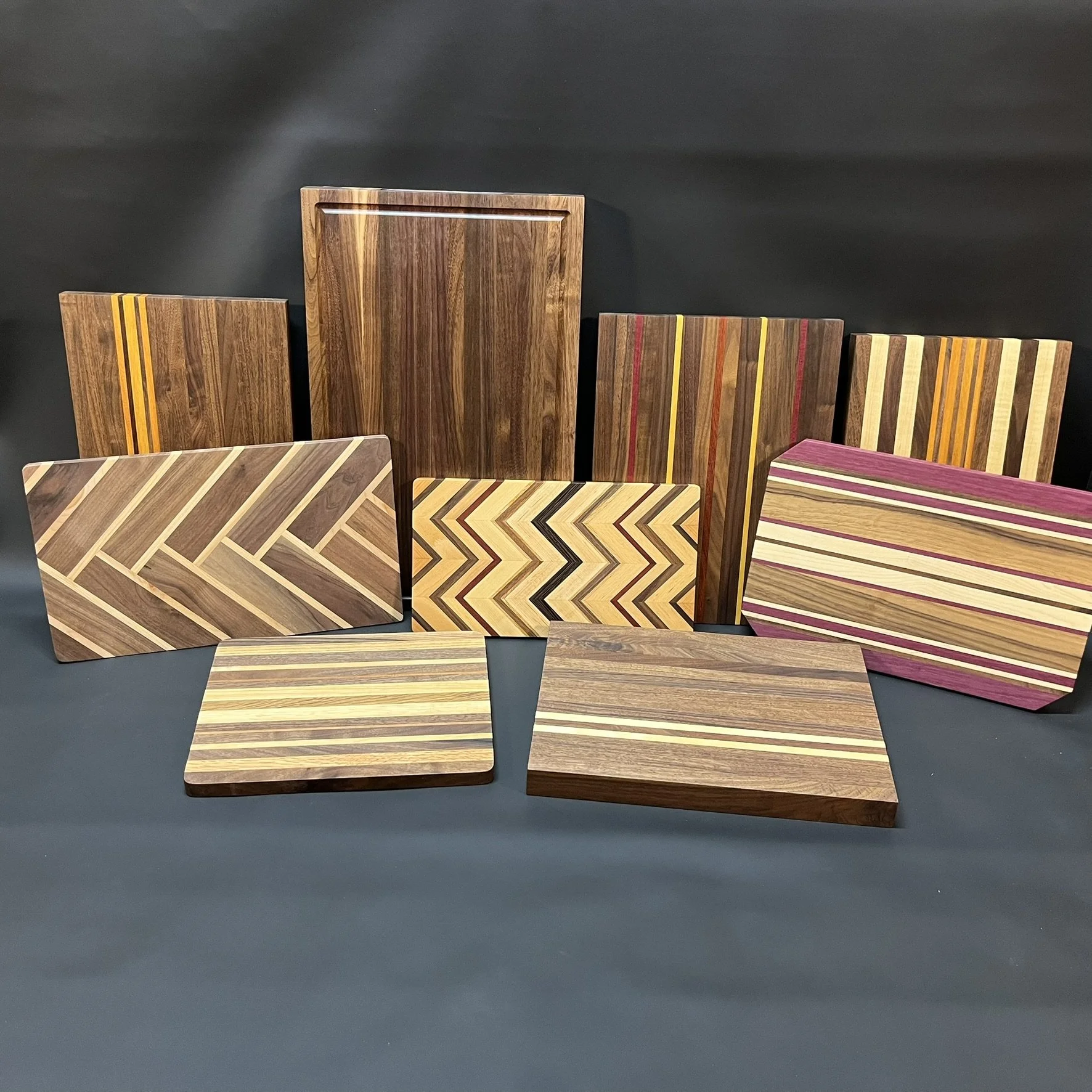 Assorted wooden cutting boards with various striped and chevron patterns, arranged on a dark surface against a black background.