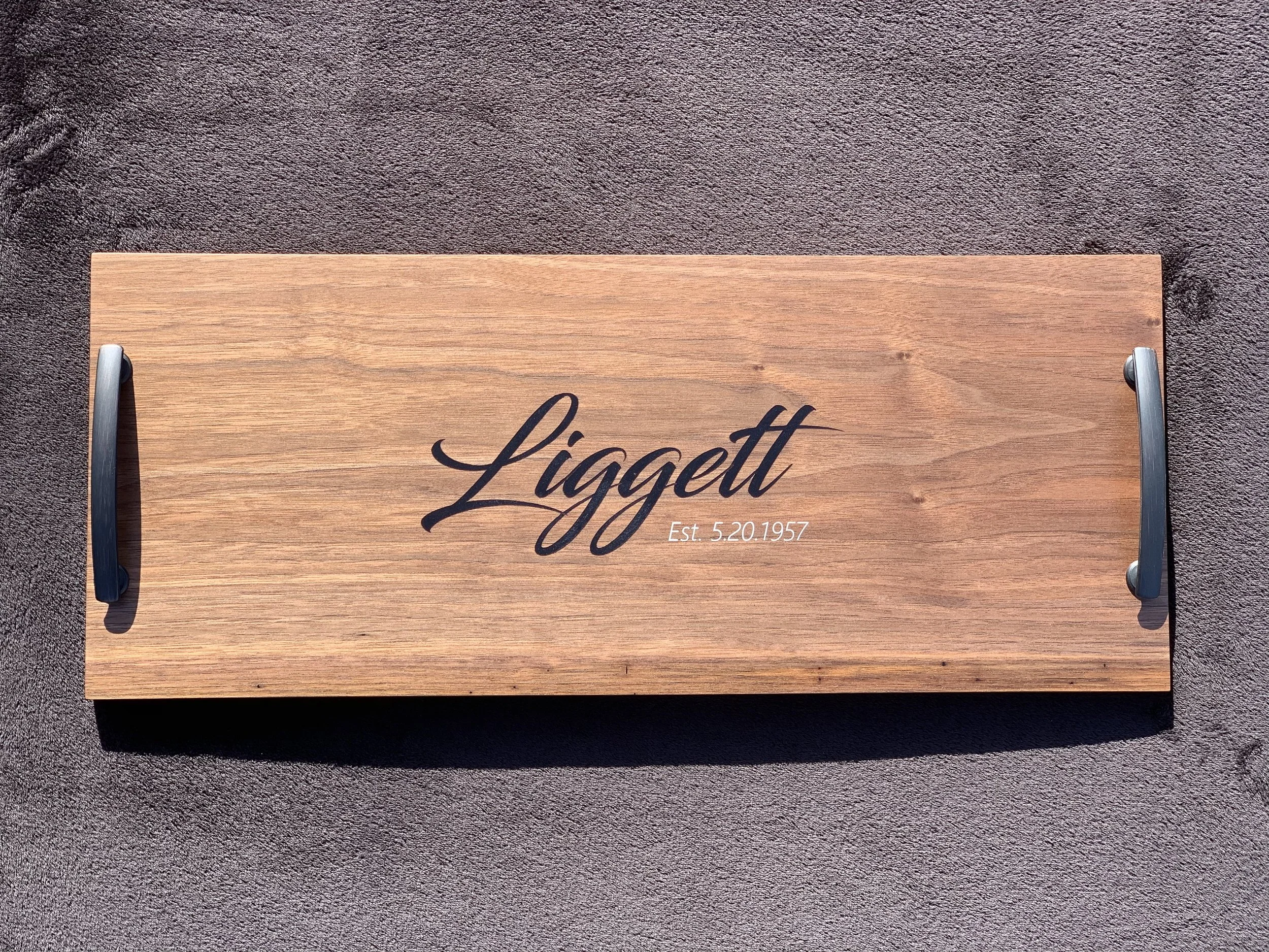 Wooden plaque with black handlebars on each side, engraved with the last name "Liggatt" and the establishment date "5.20.1957" on a textured dark surface.