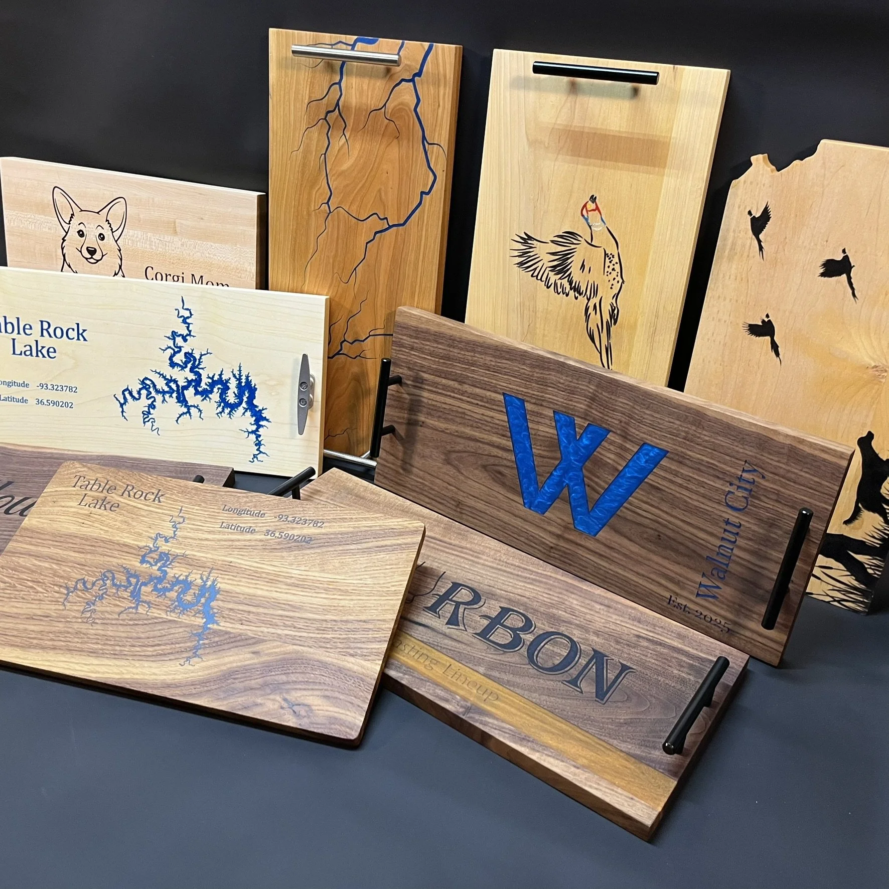 Collection of wooden cutting boards with engraved and painted designs, including a map of Table Rock Lake, a dog, a lightning bolt, a bird with a fishing lure, and initials 'W'.