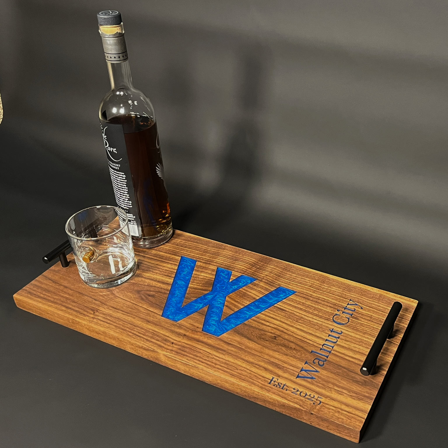 A wooden serving tray with black handles, featuring a large blue water logo and the text 'V' and 'W' in the center, 'Napalm City' and 'Est. 2025' inscribed at the bottom. On the tray, there is a glass with a small amount of amber liquid and a shot gl