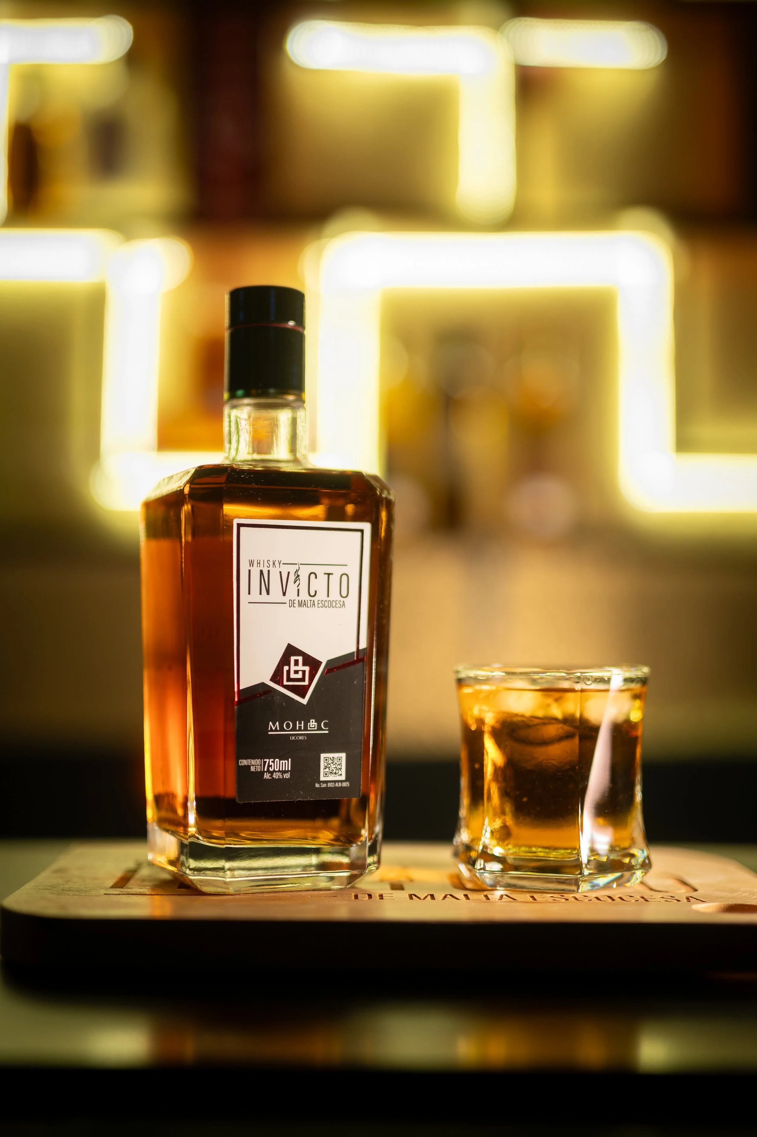 A bottle of whisky and a tumbler glass filled with whisky on a wooden surface, with a blurred background of warm light rectangles.