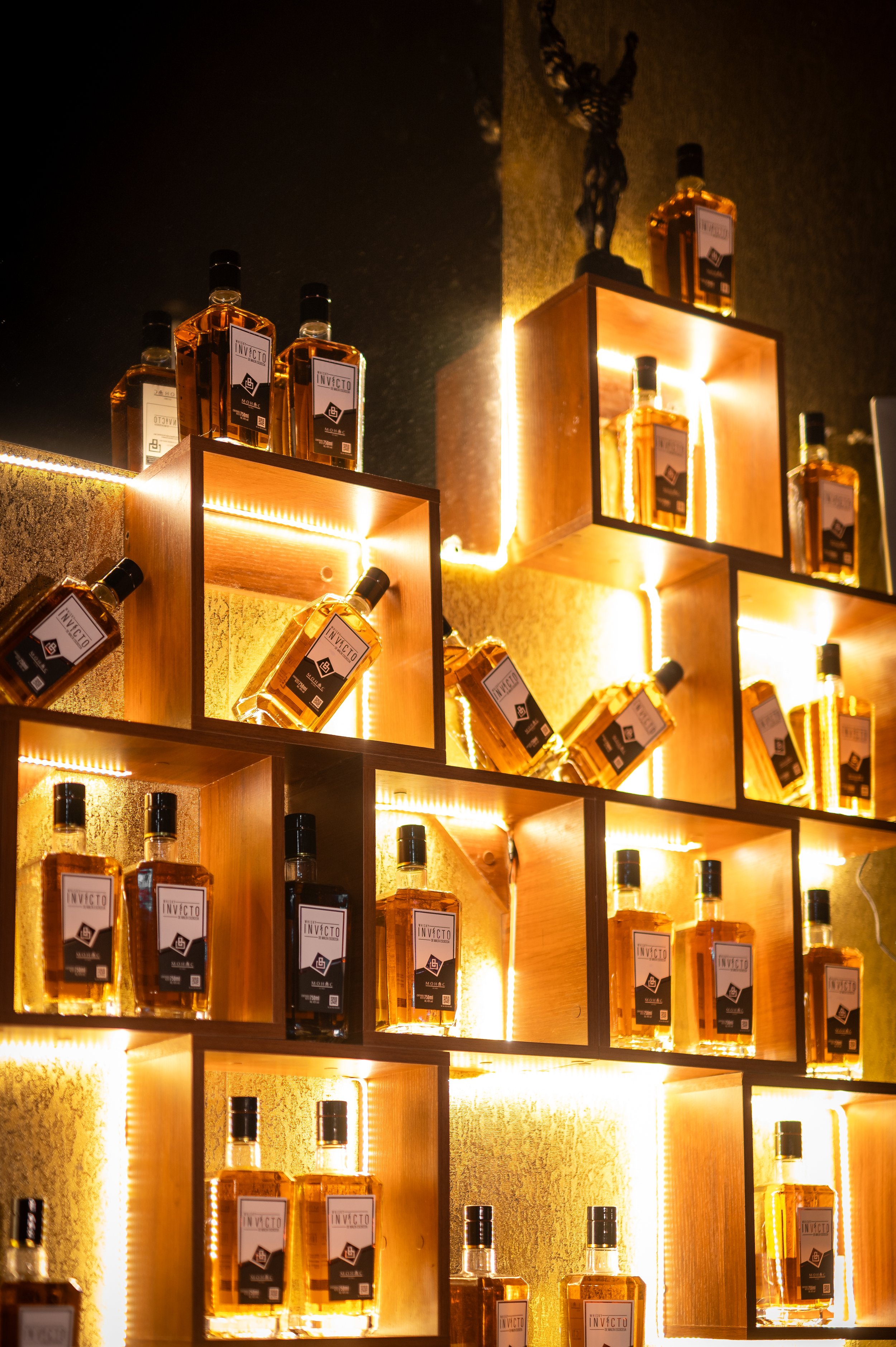 Display shelves with bottles of Invicto liquor, illuminated with warm backlighting, with a decorative sculpture on top.