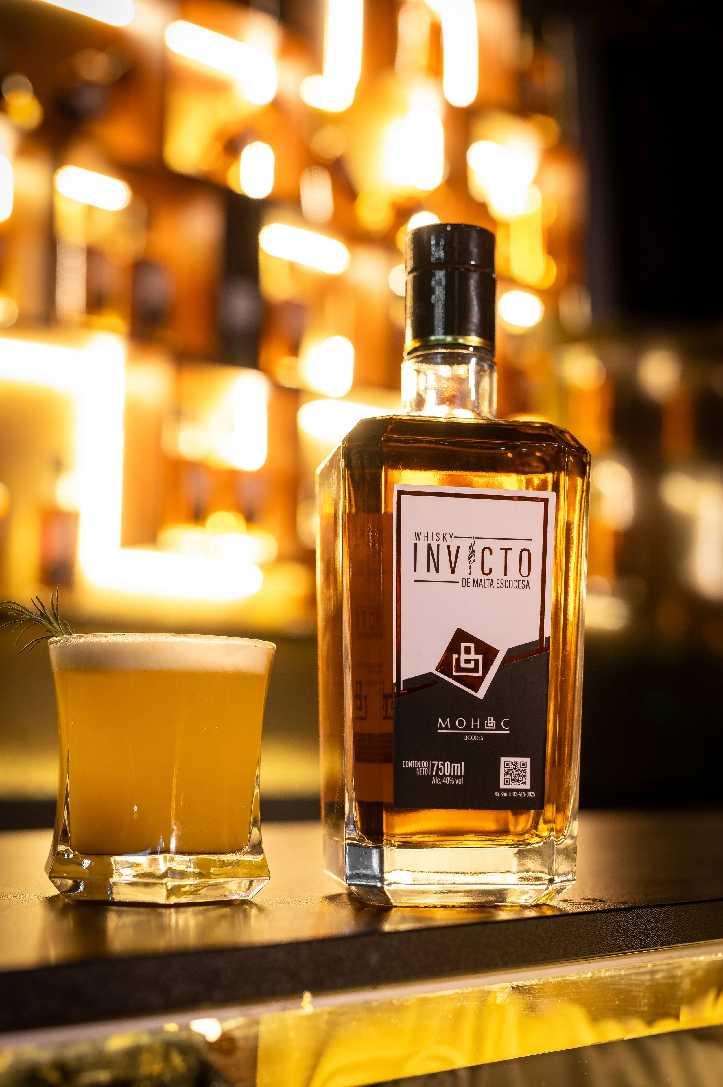 A bottle of whiskey and a cocktail glass with a garnish on a bar counter, with blurred warm lighting in the background.