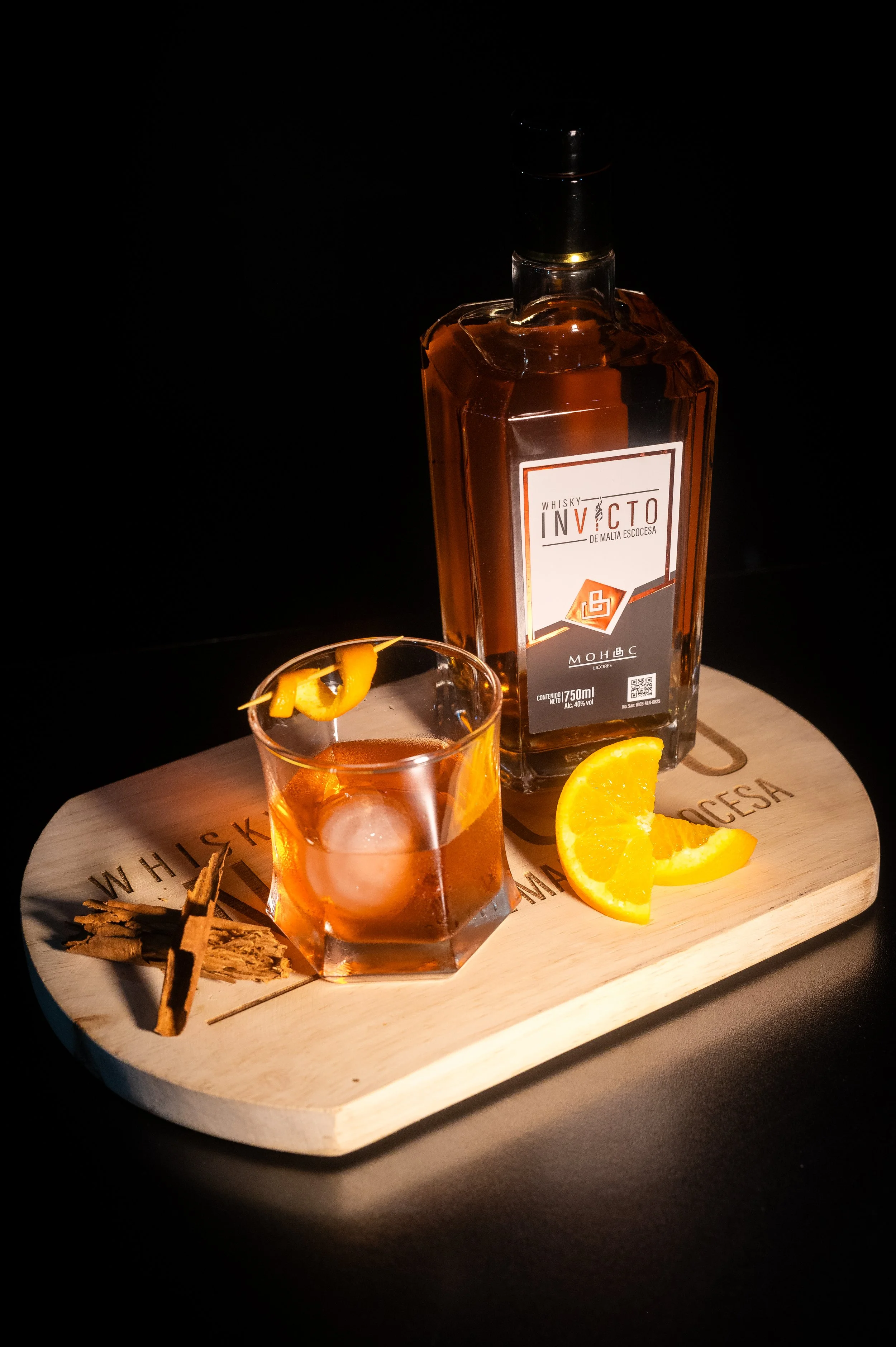 A bottle of whisky with a glass of whisky and ice, garnished with orange slices, on a wooden serving board.