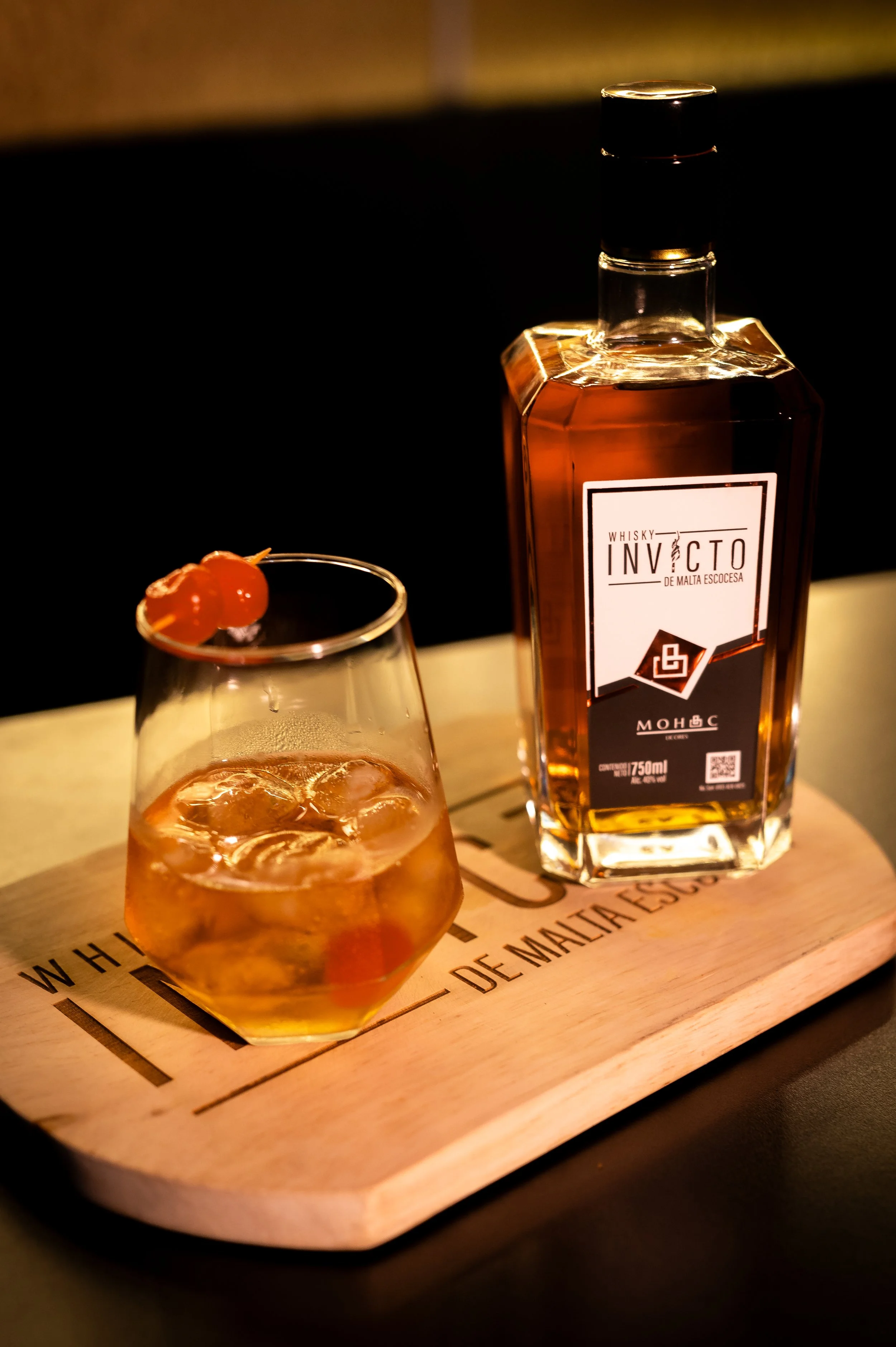 A glass of whiskey with ice and maraschino cherry garnish alongside a bottle of whiskey on a wooden serving board.