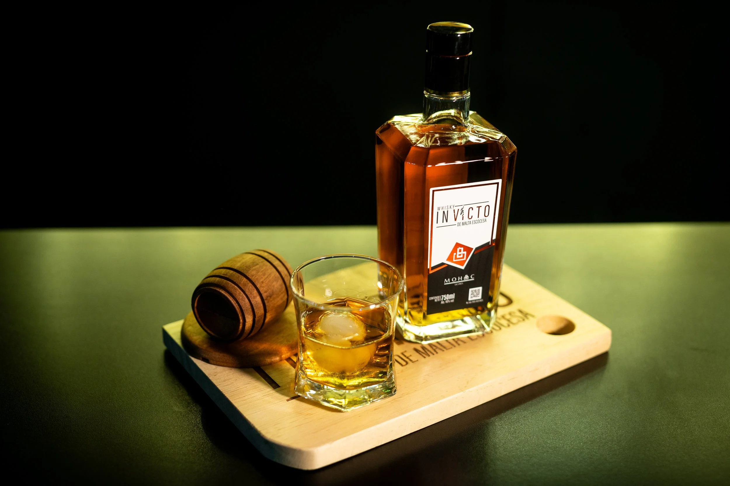 A bottle of whiskey with a glass containing whiskey and an ice cube on a wooden serving board, with a brown wooden cork beside it, on a dark background.