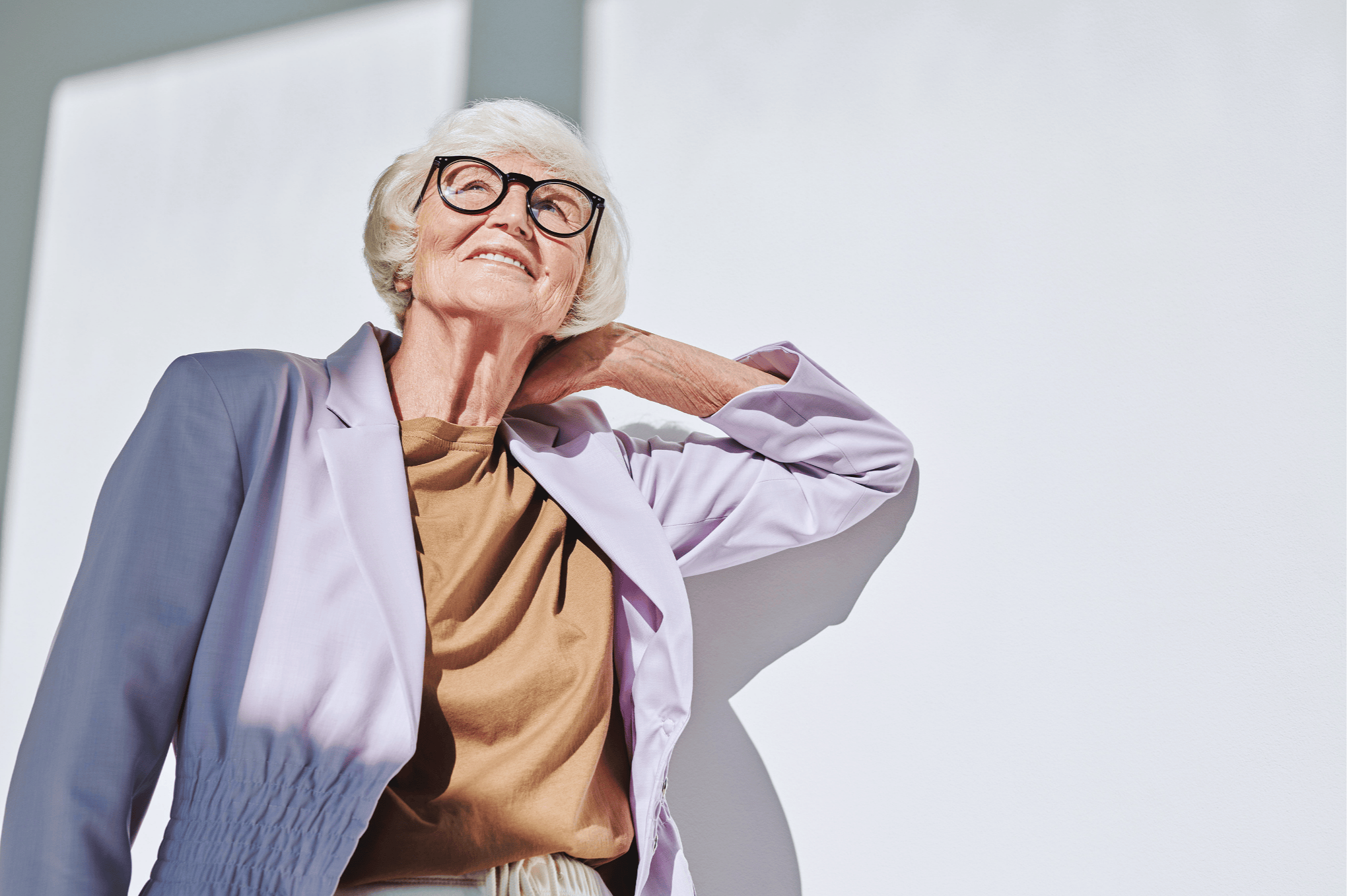 Smiling elderly woman with glasses in a tan shirt and light purple jacket outdoors on a sunny day.