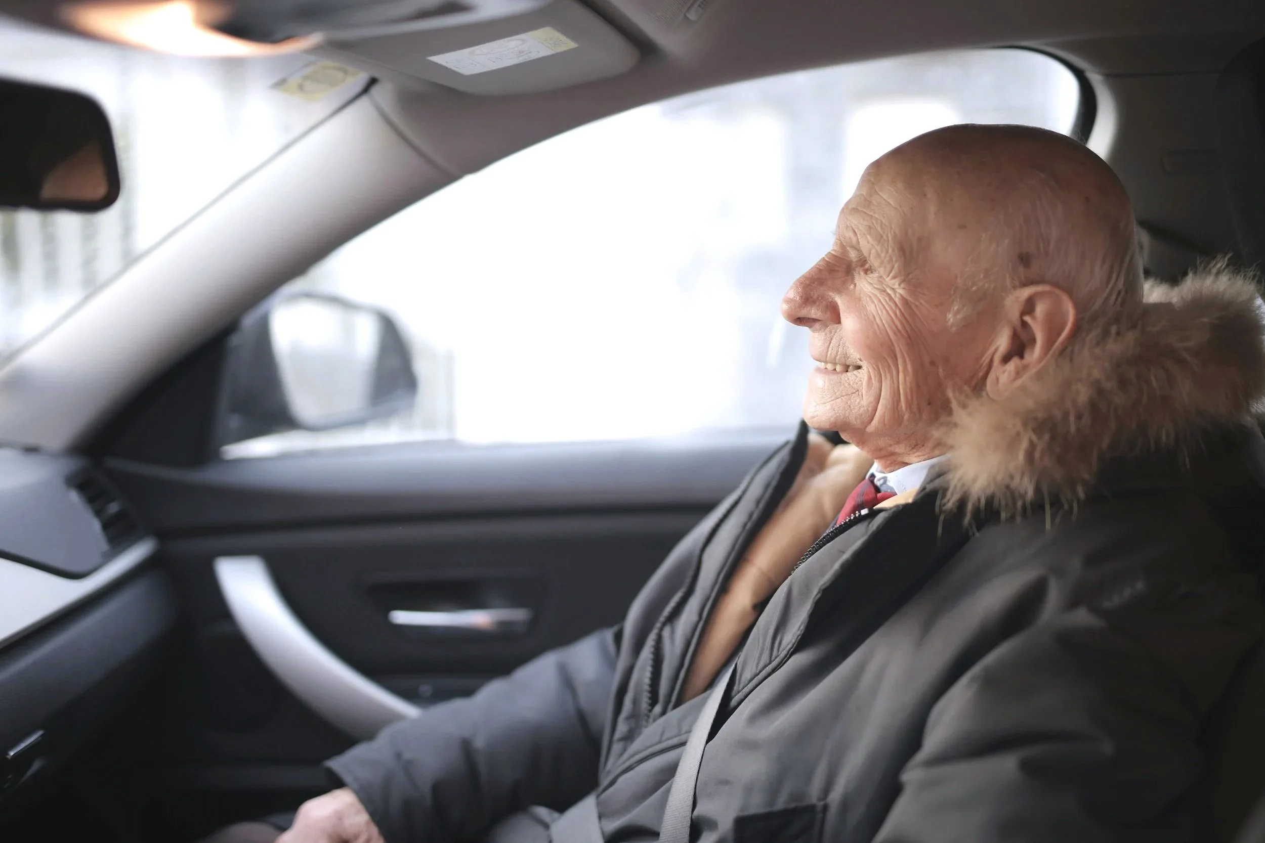 An elderly man with a bald head, smiling, sitting in the front passenger seat of a car, wearing a dark coat with a fur-lined hood.