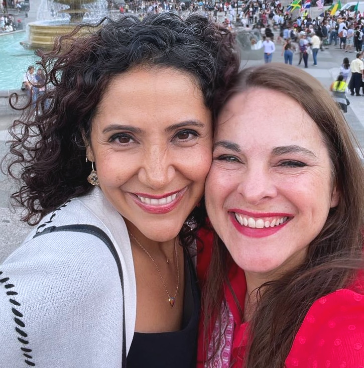 Two women smiling closely for a selfie in an outdoor park with fountains and many people in the background.