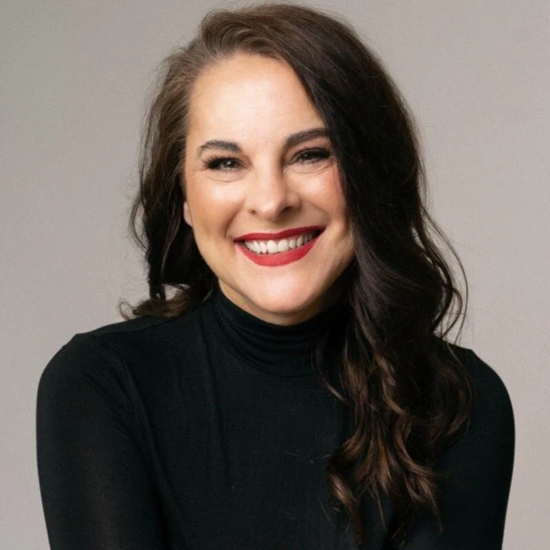A woman with long dark hair smiling, wearing a black turtleneck, against a plain neutral background.